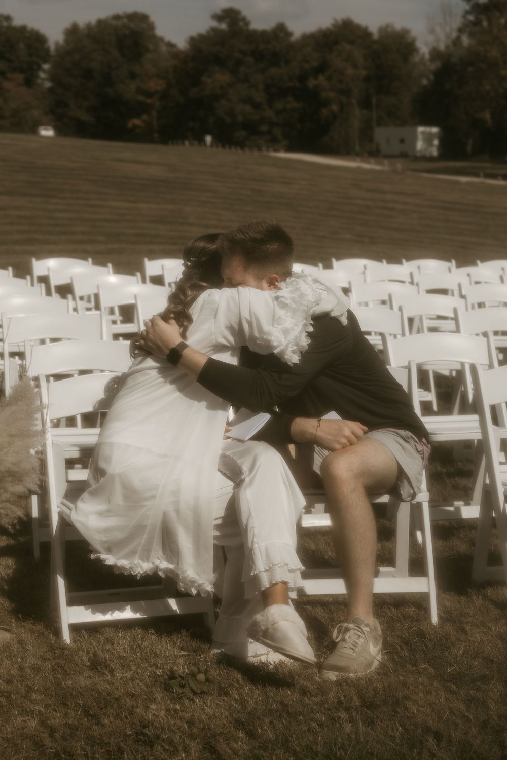Two people in white and black outfits share an intimate moment among empty chairs outdoors.