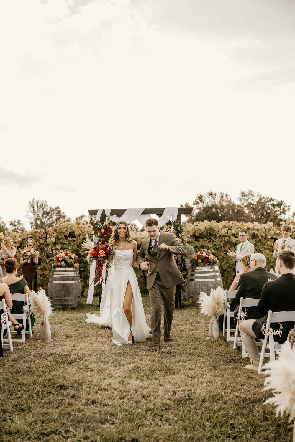 A couple walks down the aisle at an outdoor wedding ceremony surrounded by seated guests and natural scenery.