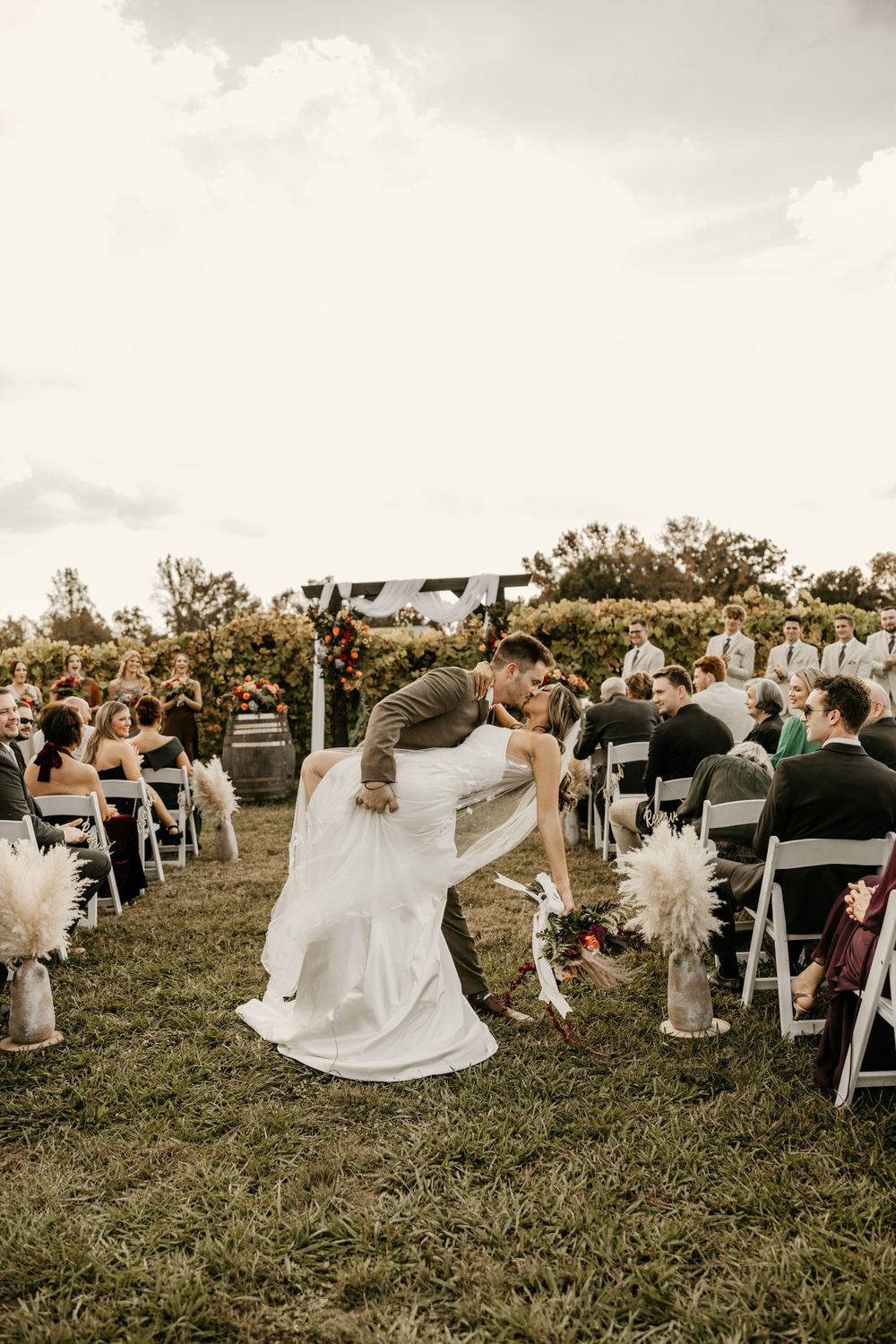 A romantic outdoor wedding ceremony on a grassy hill with white chairs and pampas grass decorations at sunset.