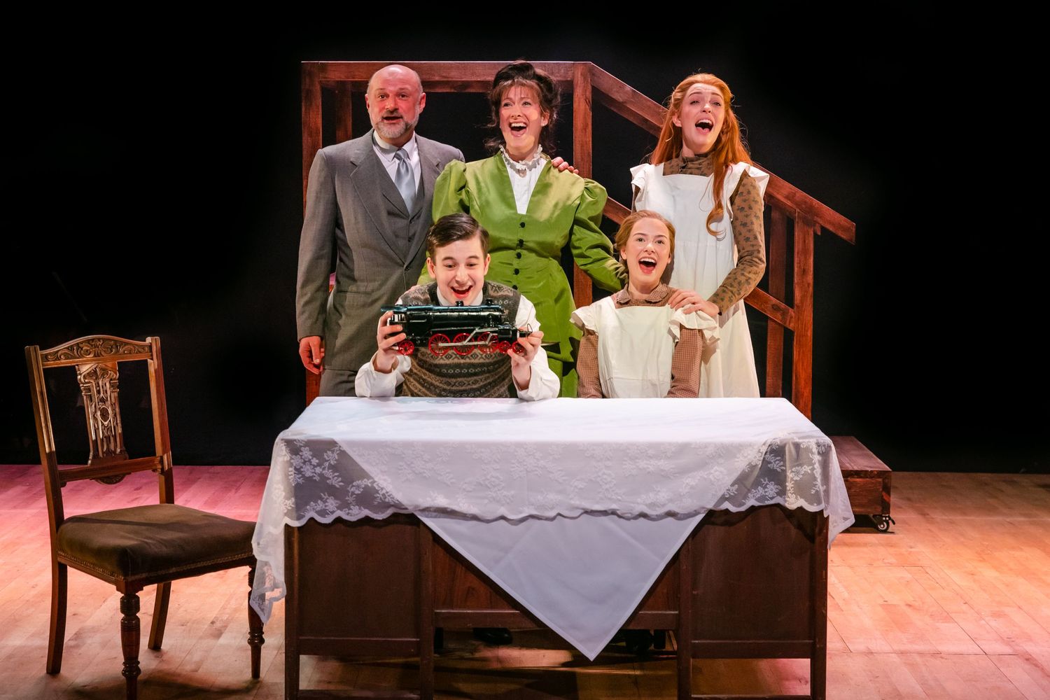 Family scene at dining table during theatrical performance with period costumes.