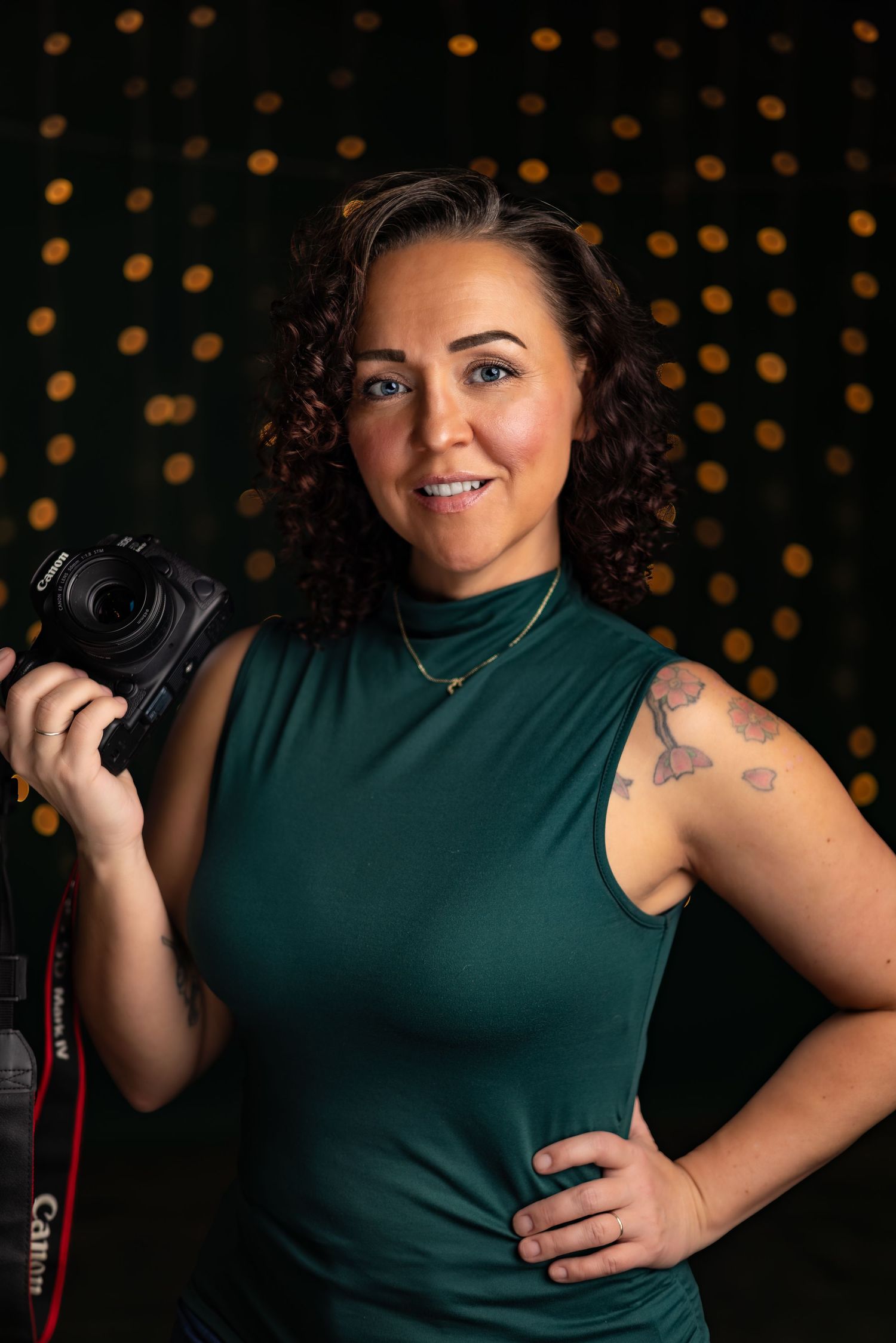 Professional photographer in dark green sleeveless top poses with camera against bokeh background.