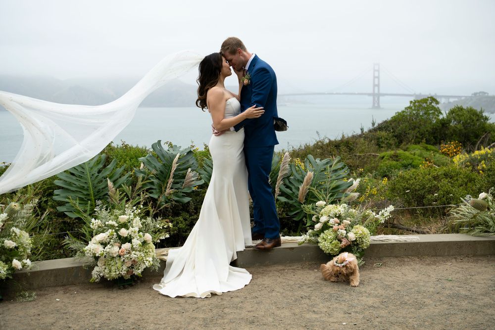 Bride and groom embracing as her veil blows in the wind with the Golden Gate Bridge in the background at Lands End in San Francisco