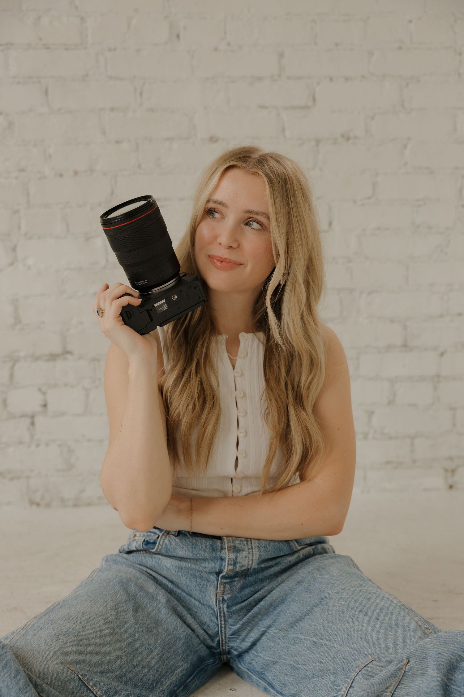 Blonde female photographer holding a camera while sitting against a white brick wall background.