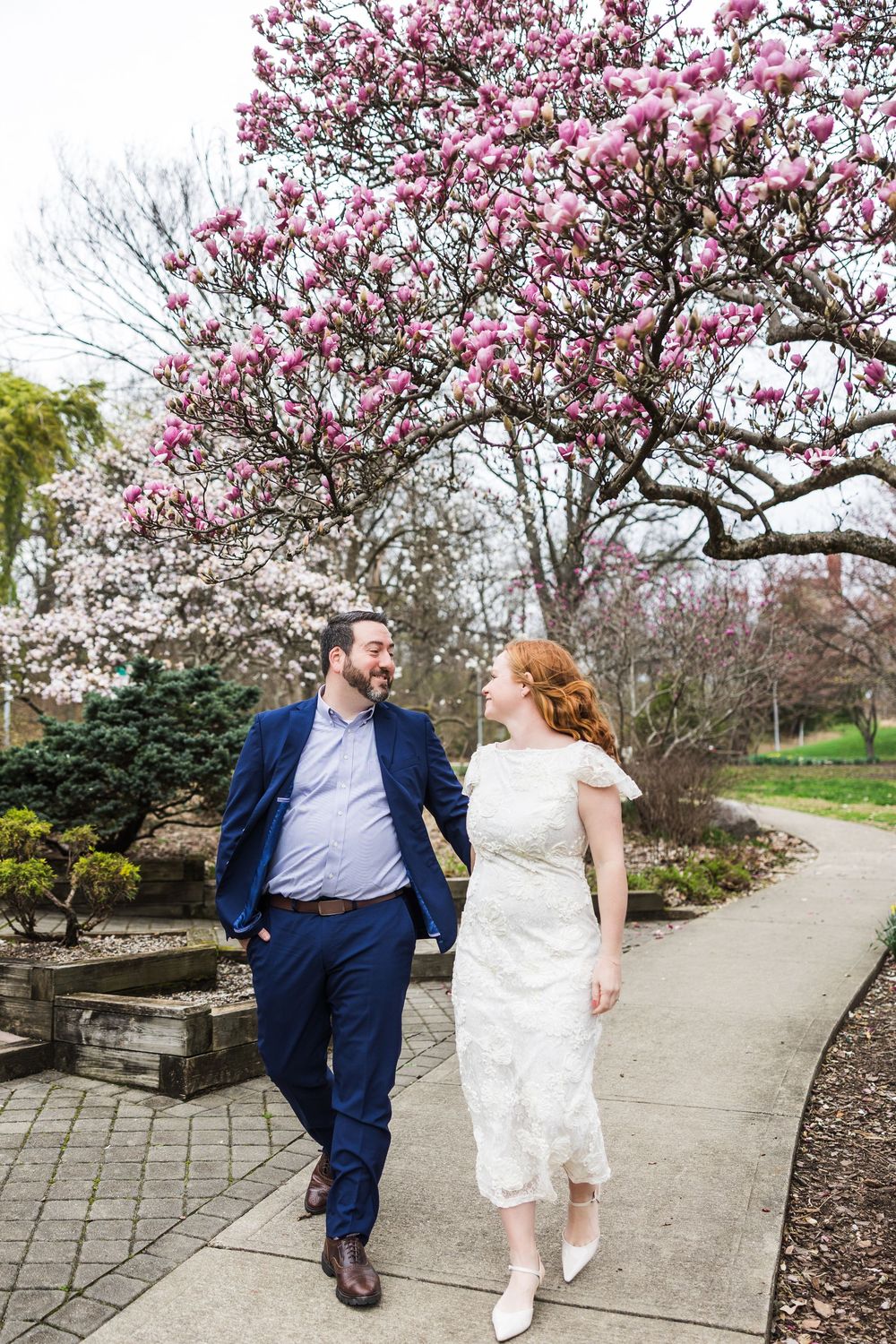 A couple walk hand in hand beneath a blooming pink magnolia tree in a garden setting.