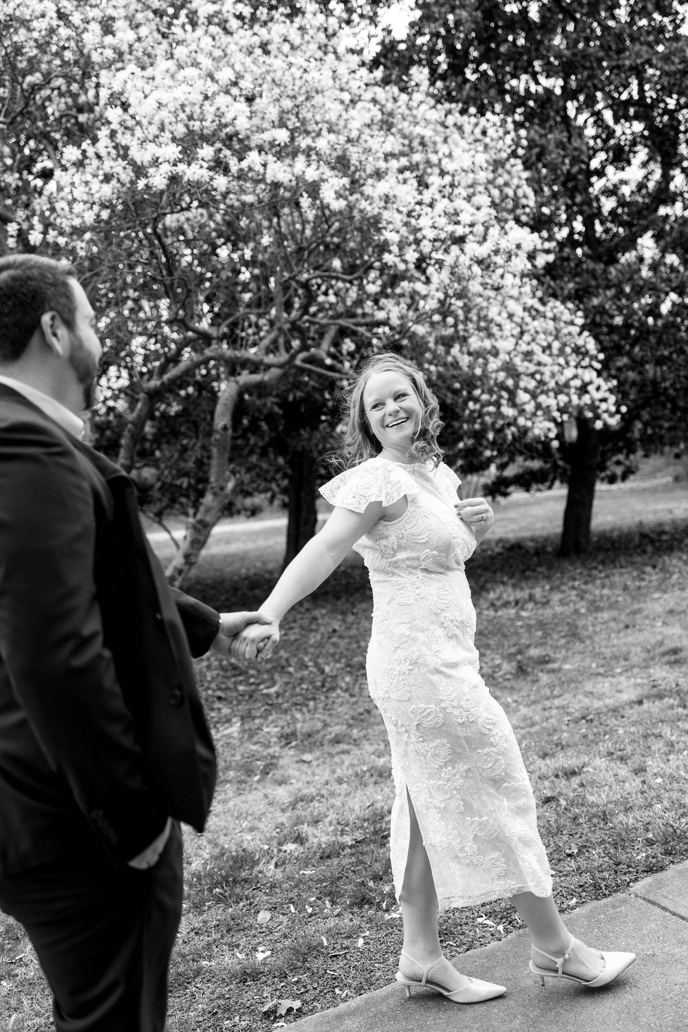 Black and white photo of a smiling woman in lace dress holding hands with man near blooming trees.