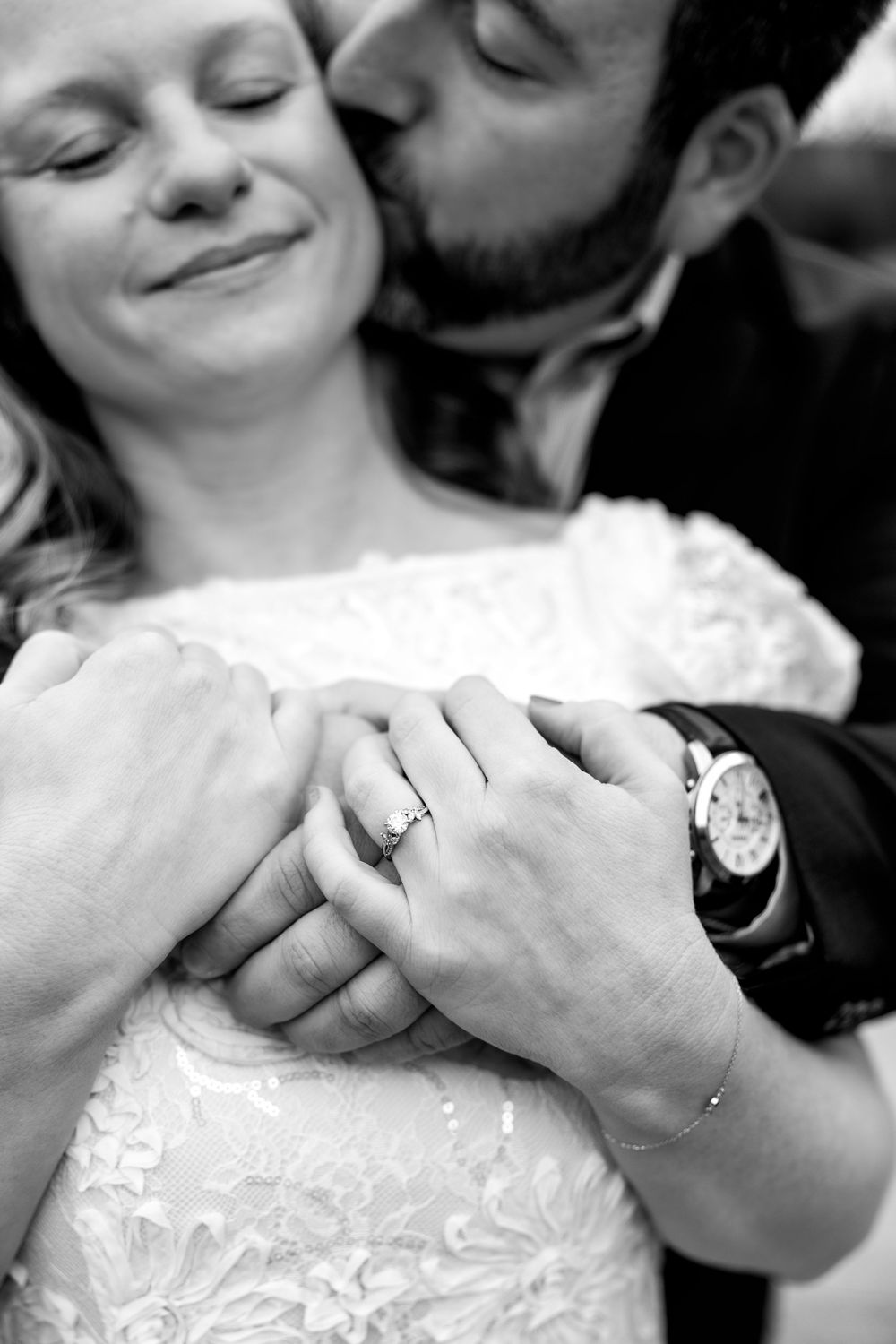 Black and white photo of a man kissing woman on cheek, hands clasped showing engagement ring and watch.