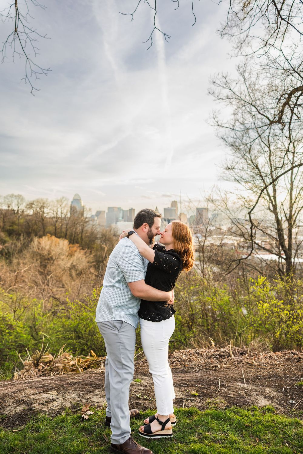 Couple sharing a romantic kiss on a hilltop with a city skyline backdrop during a spring engagement photo session.