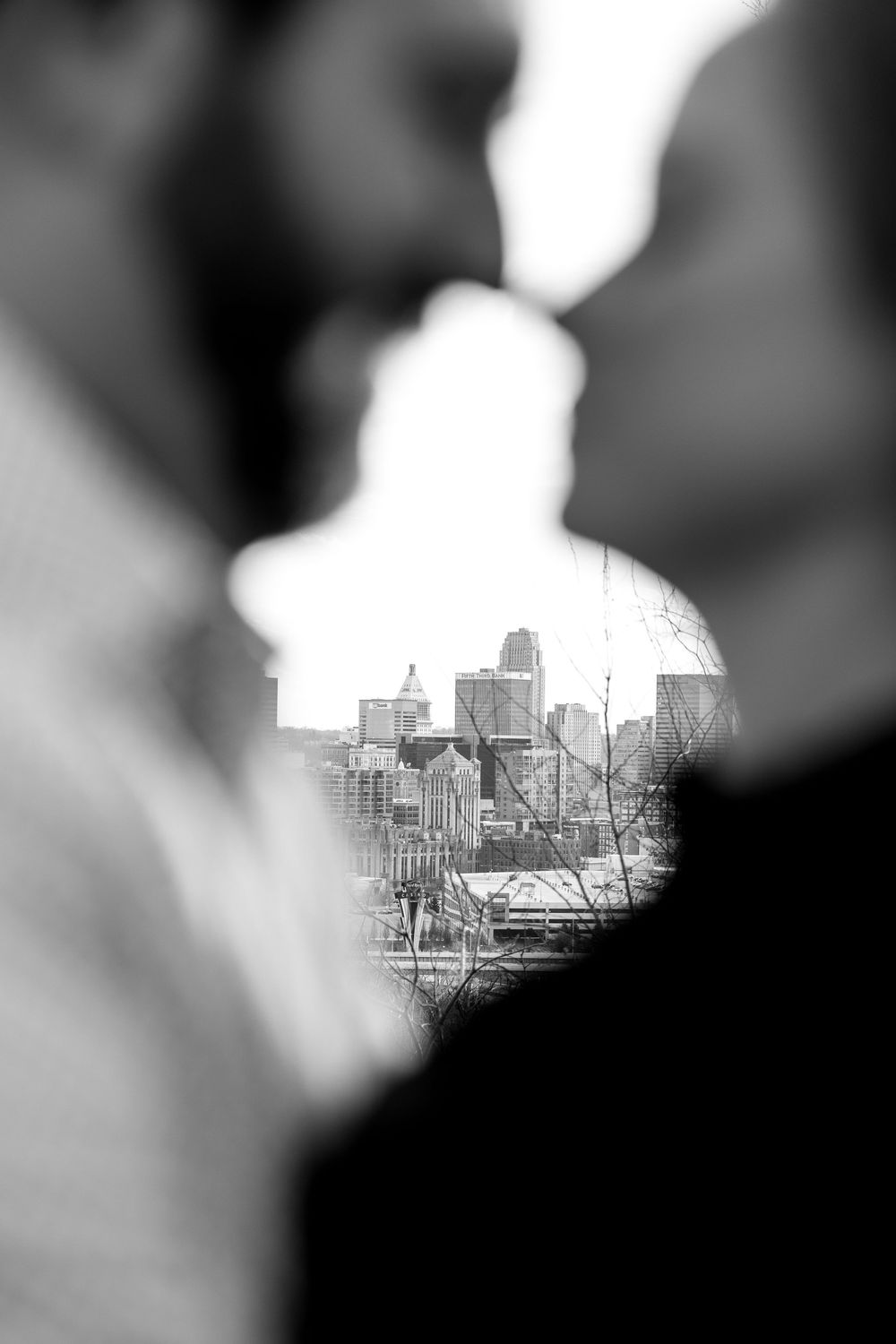 Black and white photo of couple nearly kissing with city skyline visible between them in soft focus background.