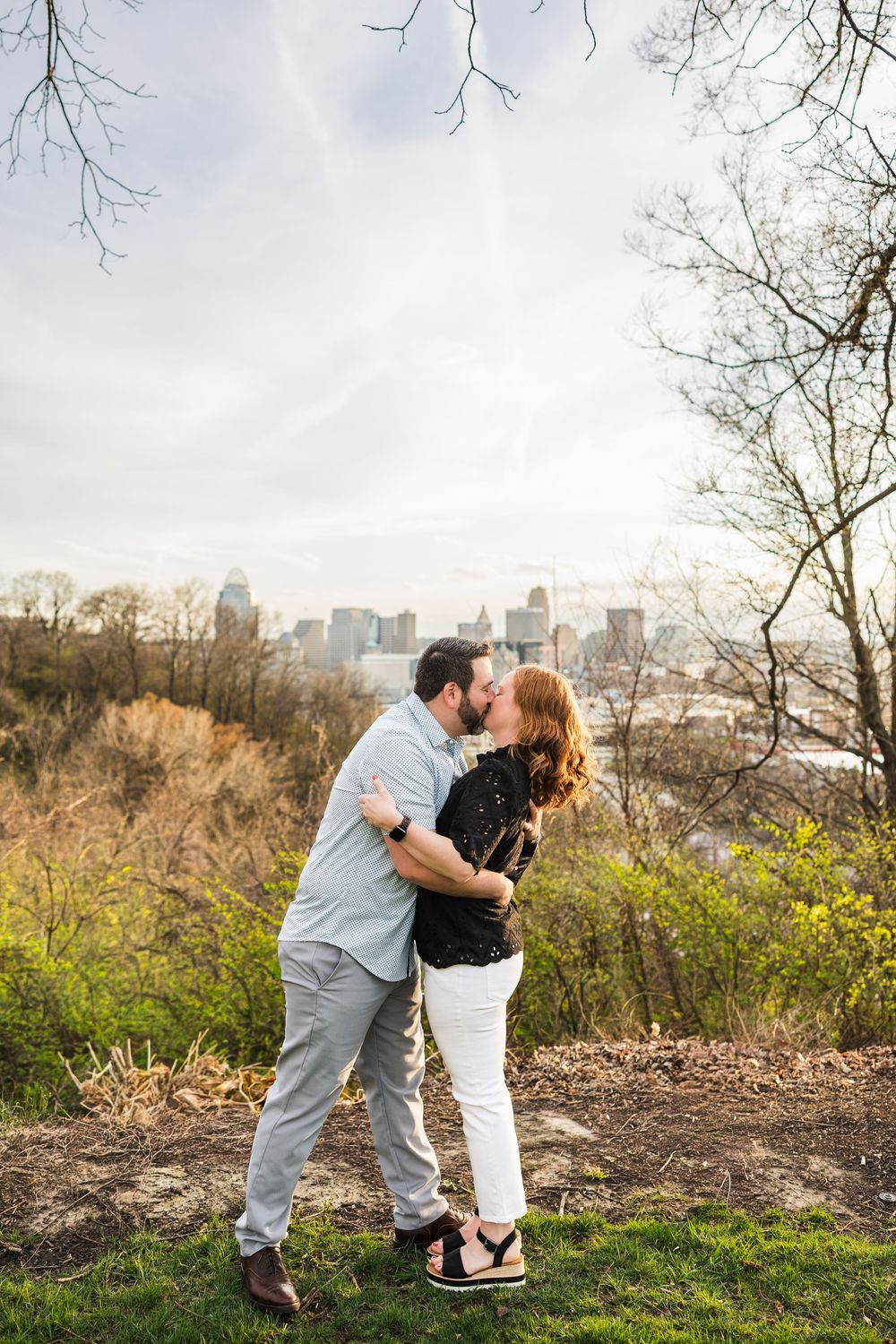 Couple sharing a romantic kiss on a hilltop with a city skyline backdrop during a spring engagement photo session.