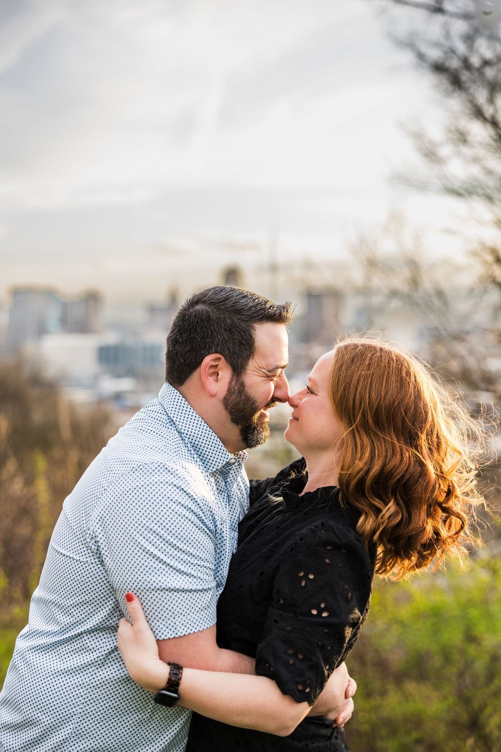 A couple shares a romantic nose-to-nose moment outdoors, with a city skyline visible in the background.