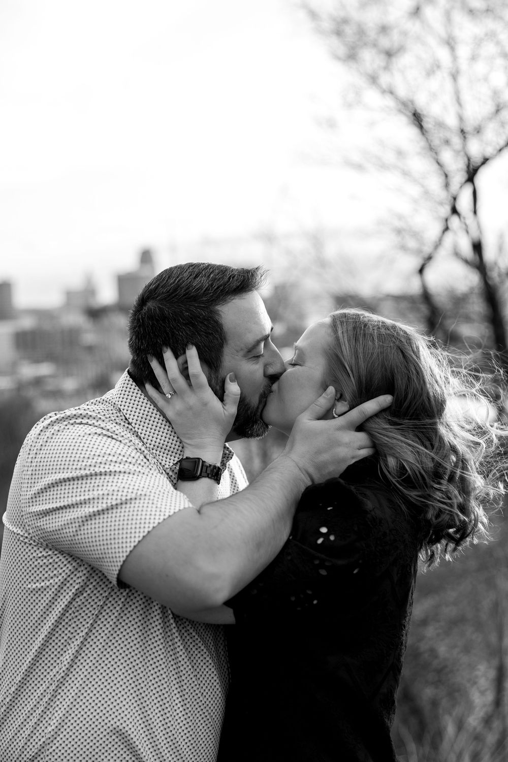 Black and white photo of a couple sharing a passionate kiss outdoors with a city skyline in the background.