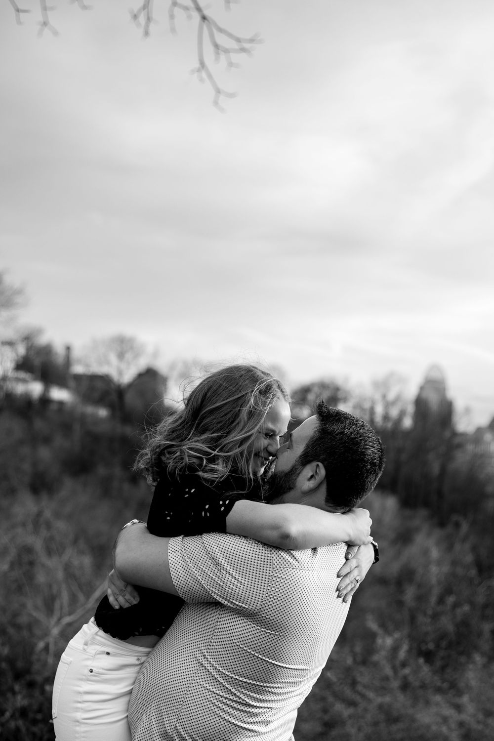 Black and white photo of a couple embracing and kissing outdoors, surrounded by trees and cloudy sky.