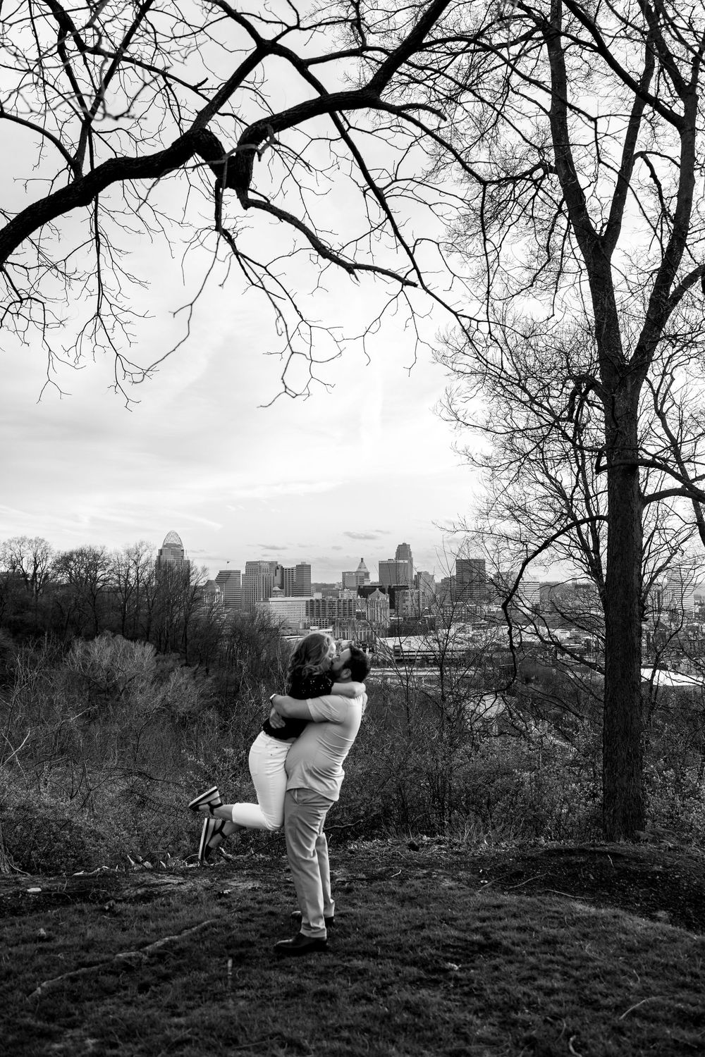 Black and white photo of couple embracing with city skyline backdrop and bare winter trees.