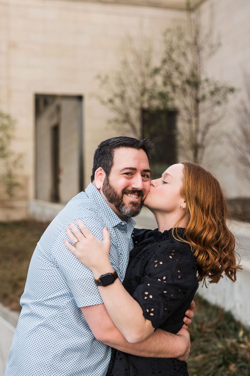 Smiling bearded man embraces redhead woman kissing his cheek outdoors near stone building.