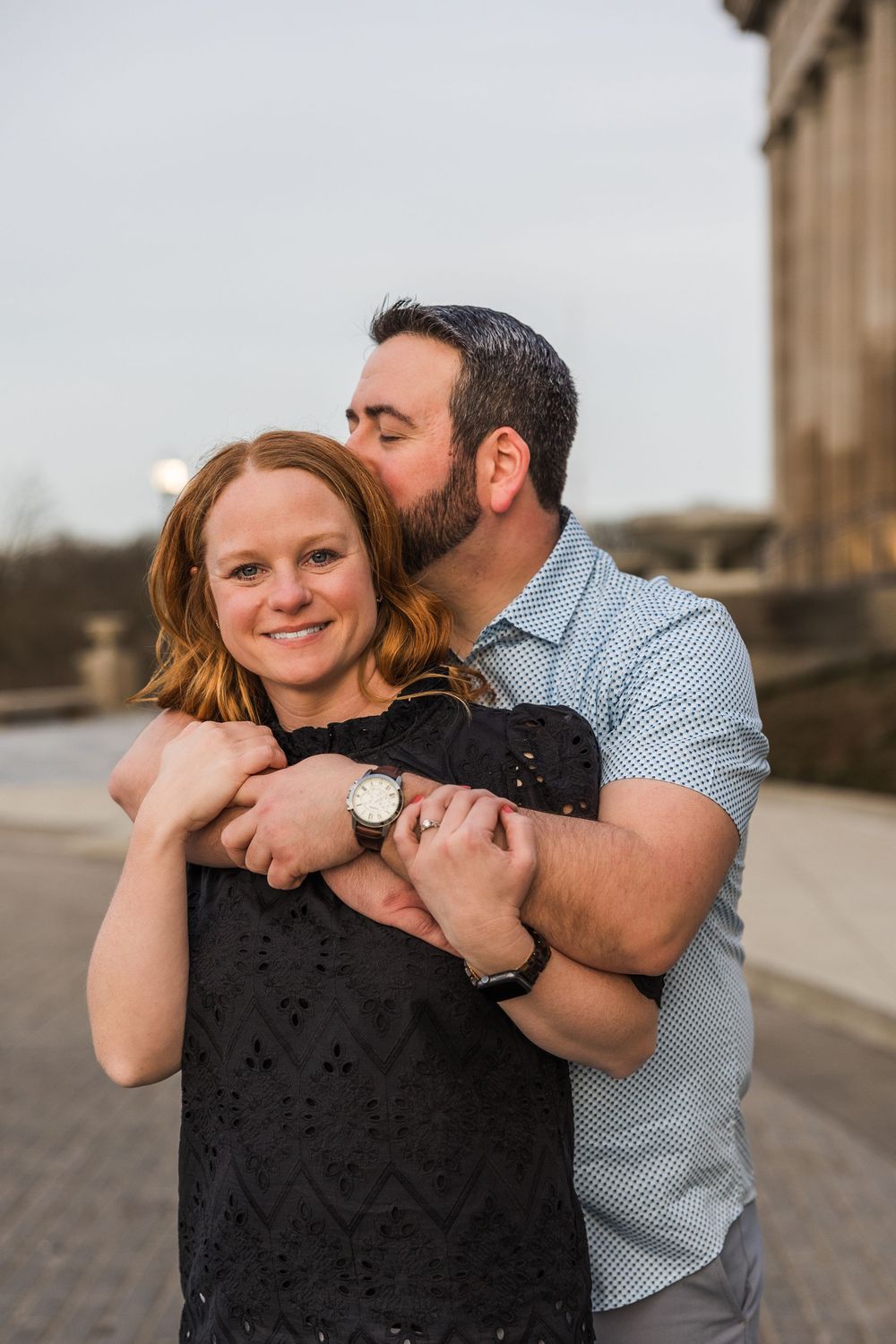 Bearded man kisses smiling redheaded woman on cheek while embracing her on urban steps.