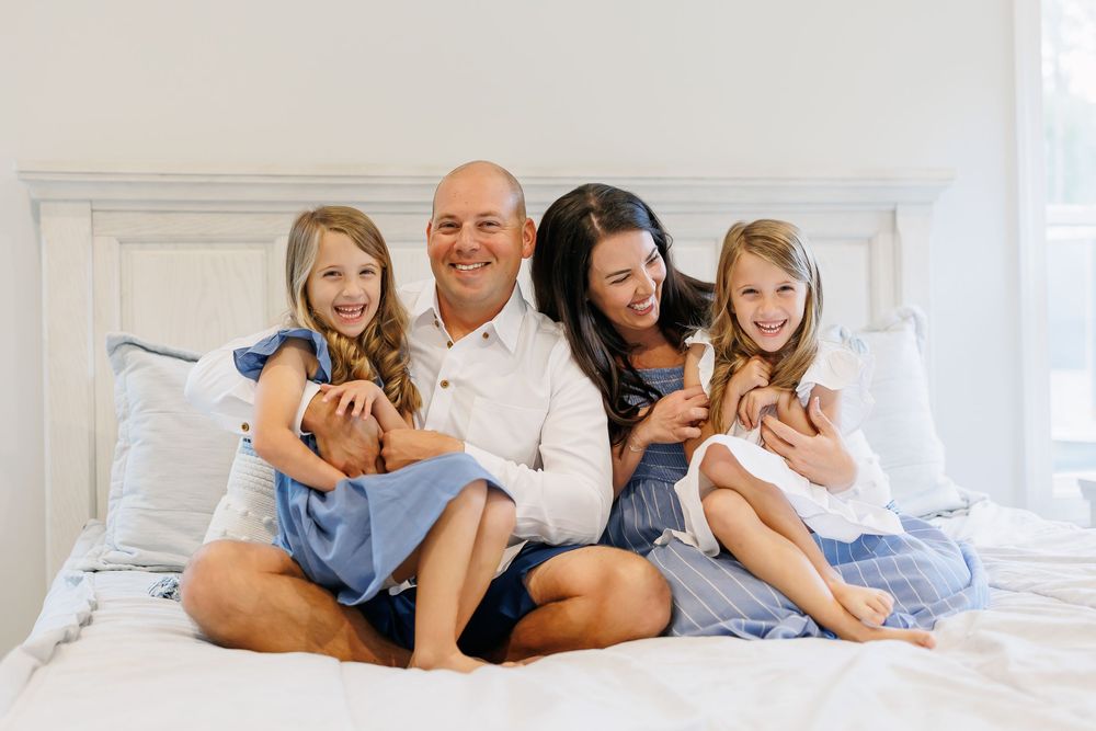 A family lounges together on a white bed wearing casual clothes in a bright, airy bedroom.