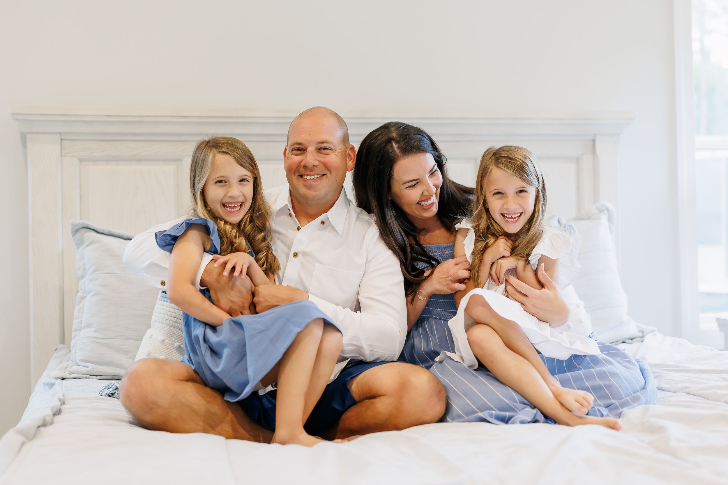 A family lounges together on a white bed wearing casual clothes in a bright, airy bedroom.