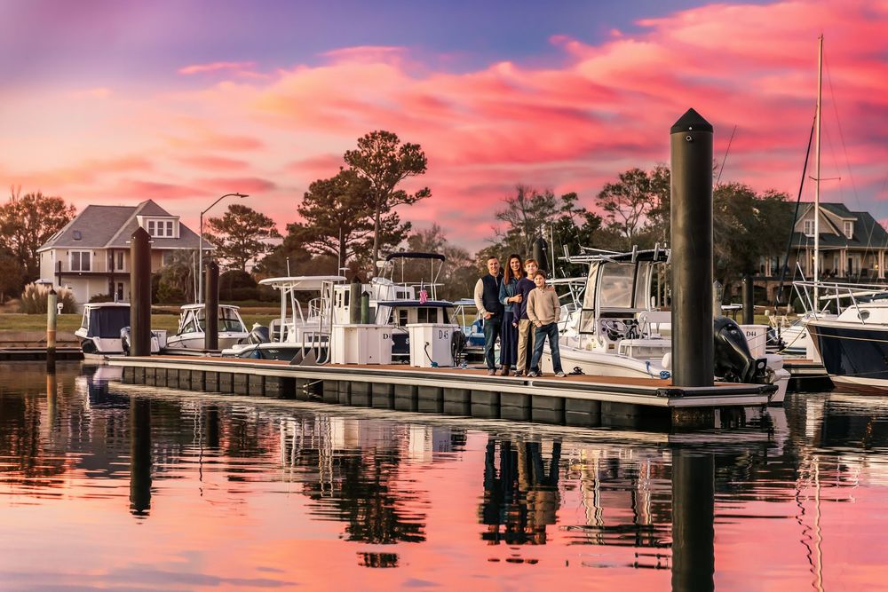Boats docked at a marina during vibrant sunset with coastal homes and pink-orange sky reflecting on calm waters.