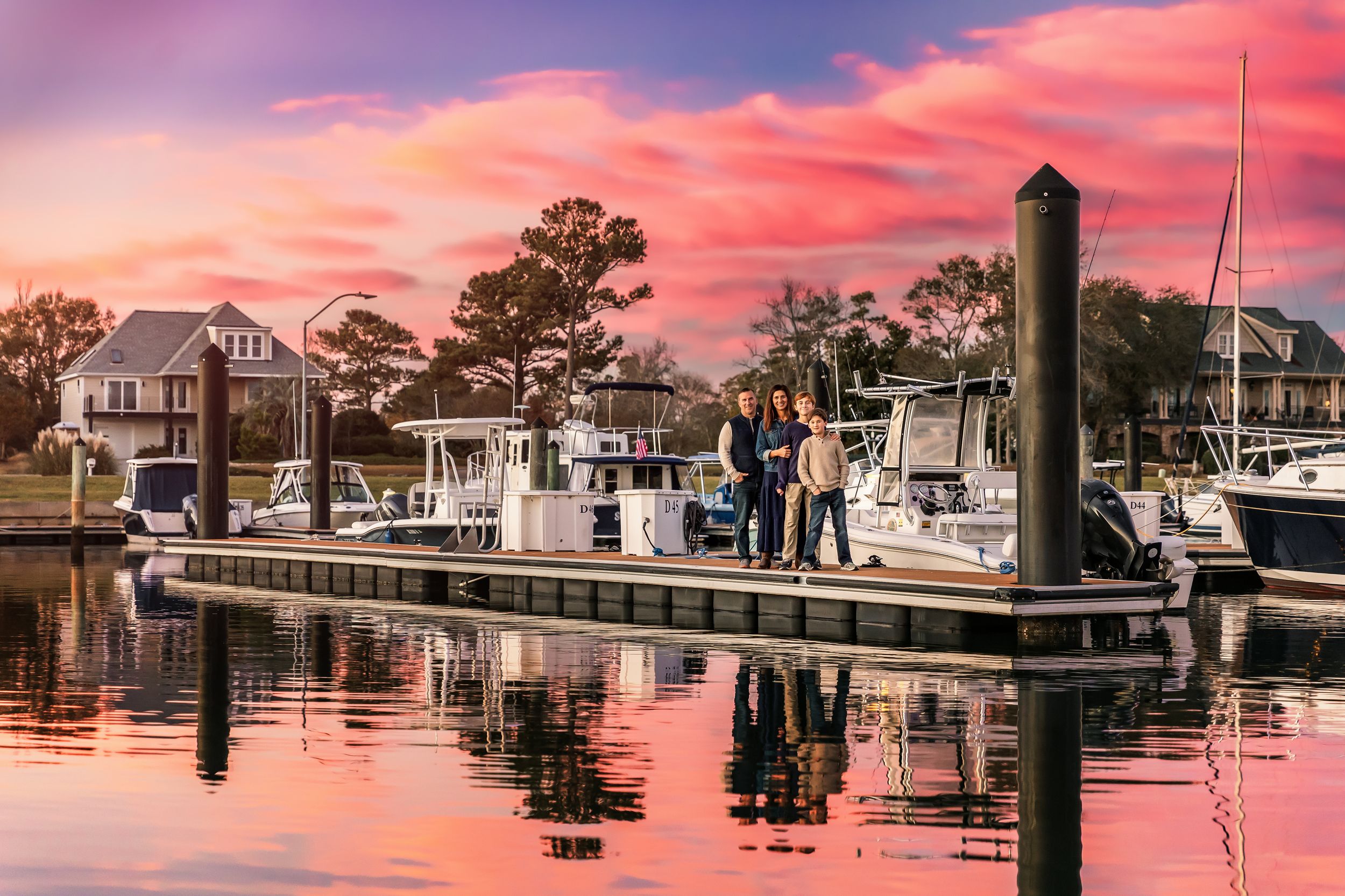 Boats docked at a marina during vibrant sunset with coastal homes and pink-orange sky reflecting on calm waters.