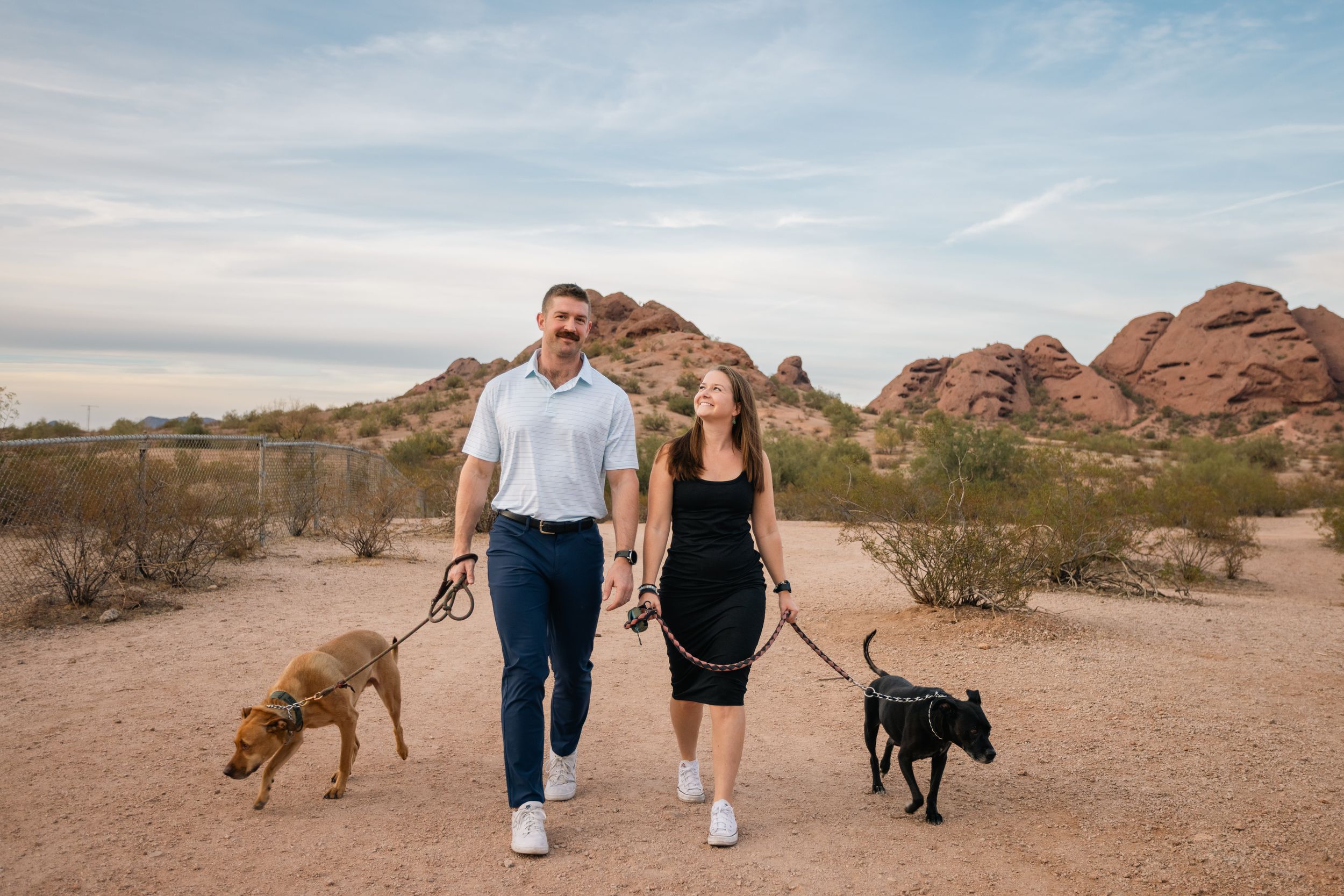 Couple walking two dogs on leashes in Arizona desert landscape with red rock formations and blue sky.