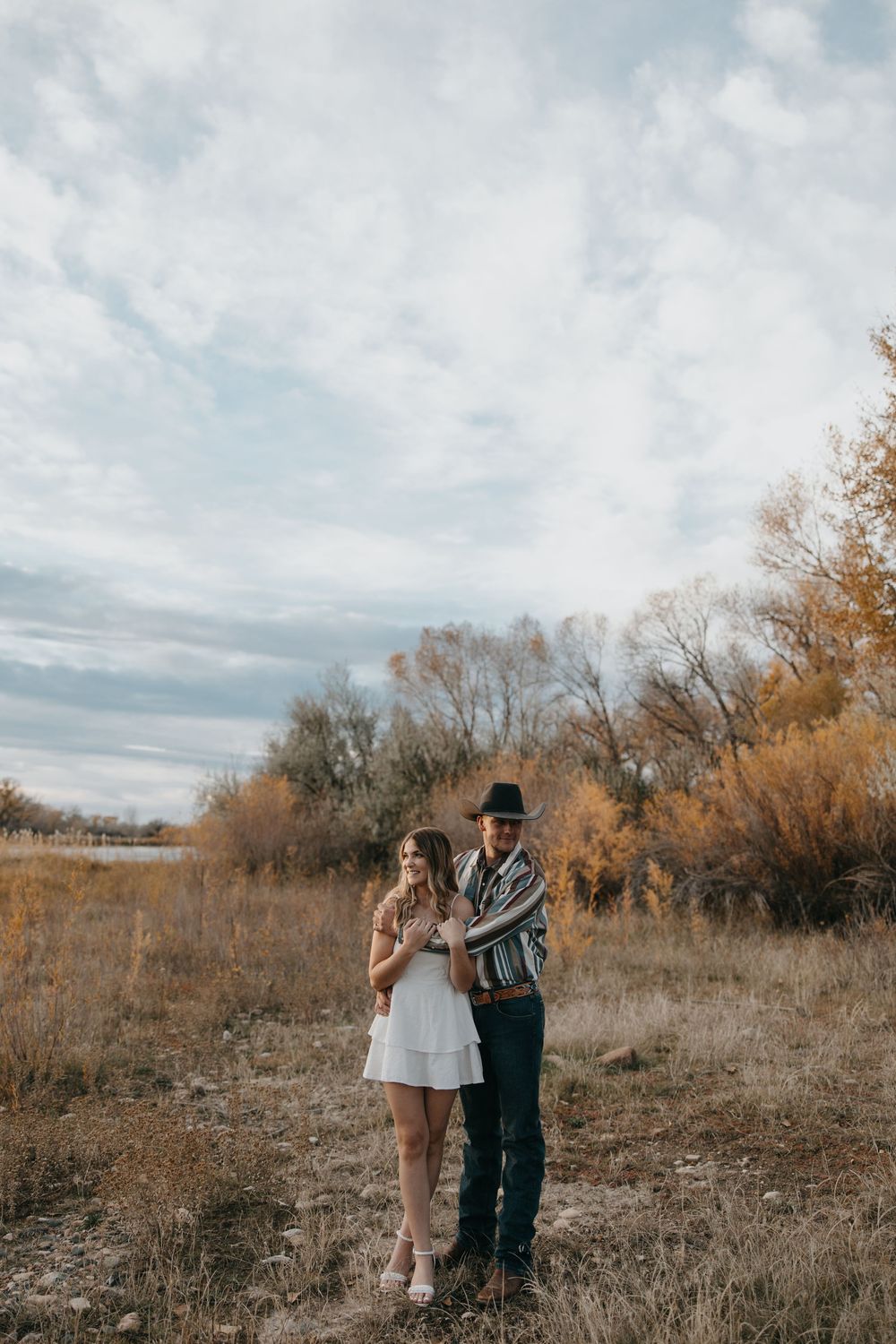 A couple stands together in a rustic autumn field with dried grass and bare trees under cloudy skies.