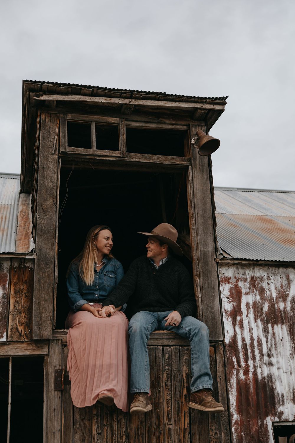A couple sits together in a rustic doorway of an old barn wearing casual clothing and a brown hat.