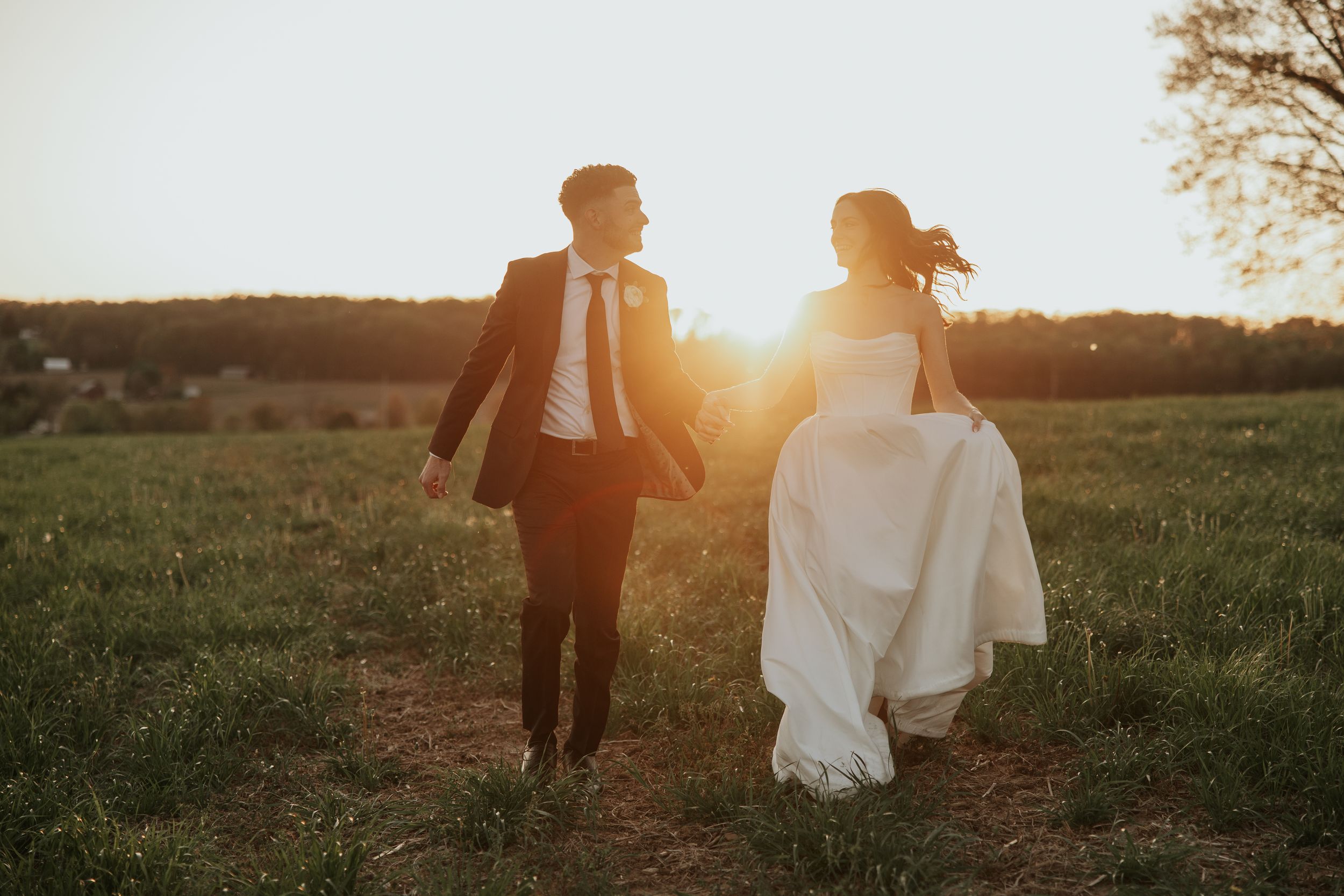 A wedding couple runs hand in hand through a sun-lit meadow at golden hour while their formal attire flows in the breeze.