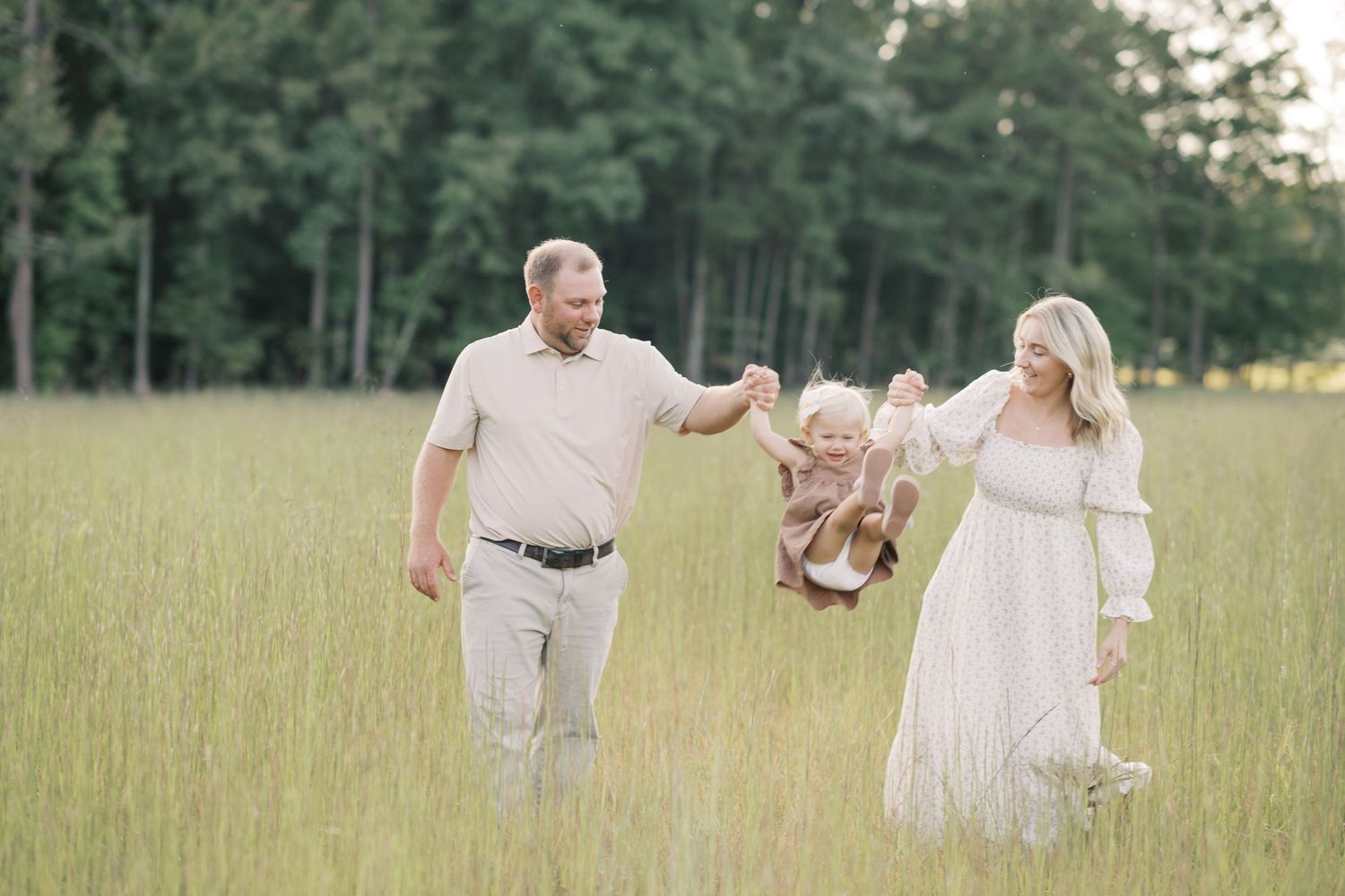 Happy family swinging their toddler child in a lush green field during a family photo session.