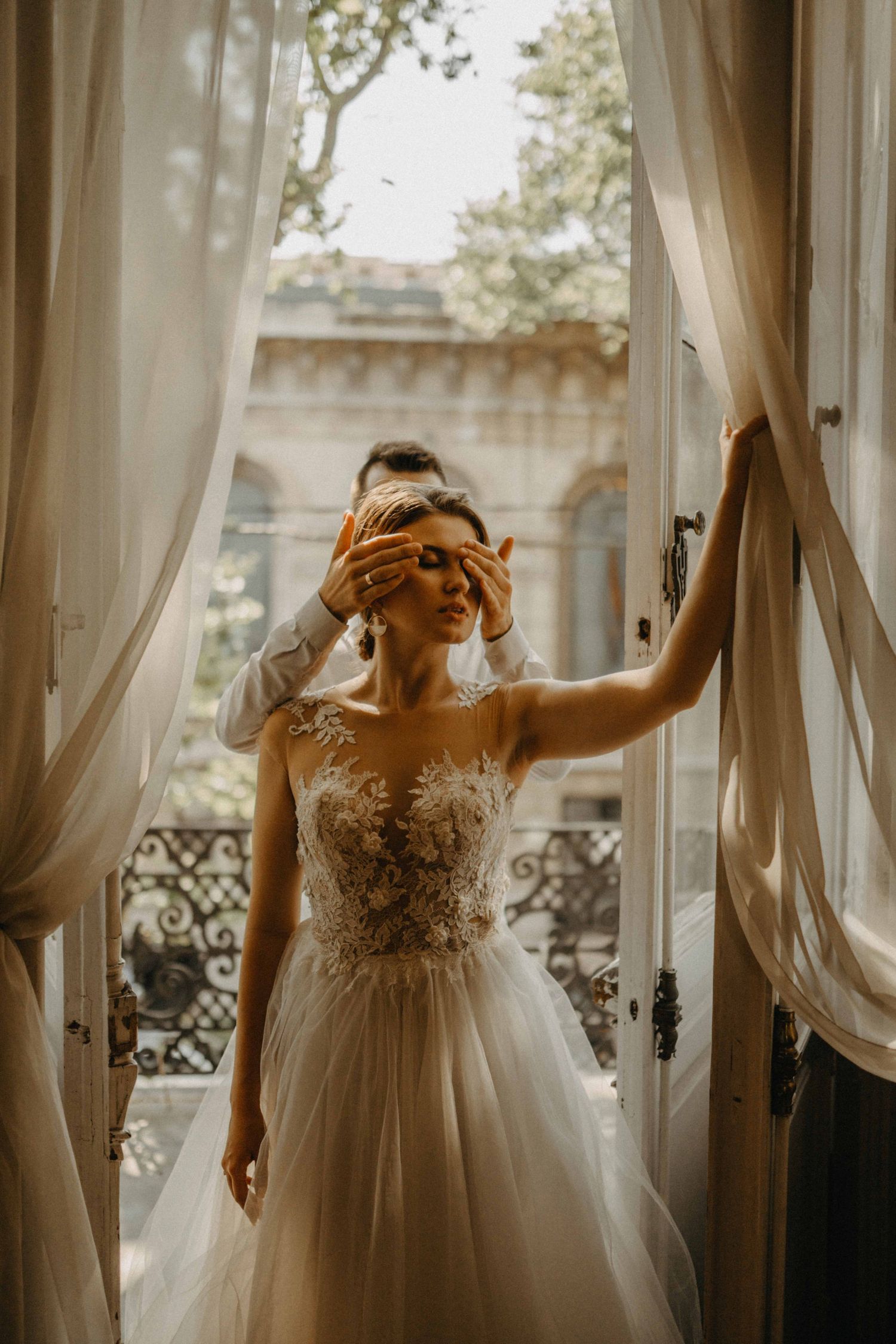 Bride in floral lace wedding gown stands in open French doors while groom covers her eyes.