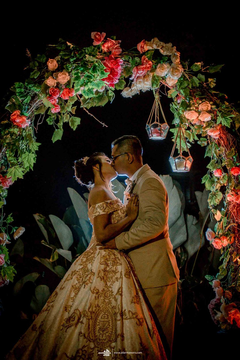 Romantic wedding scene under a floral arch with glowing lanterns and red roses against a dark background.