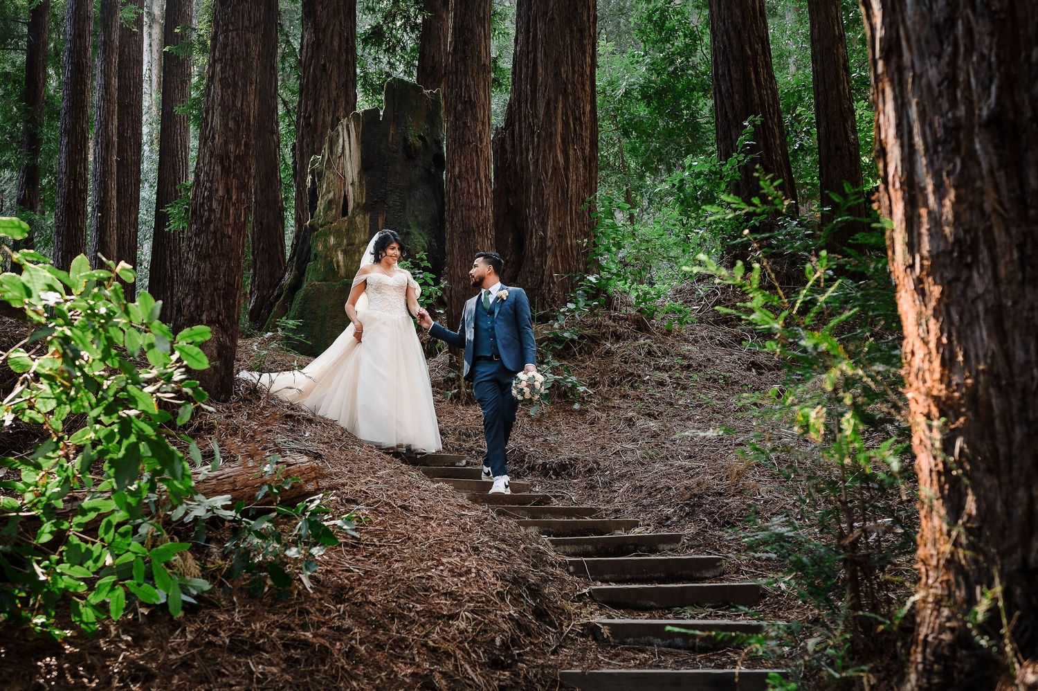 A couple walks hand in hand along rustic forest steps surrounded by towering redwood trees.