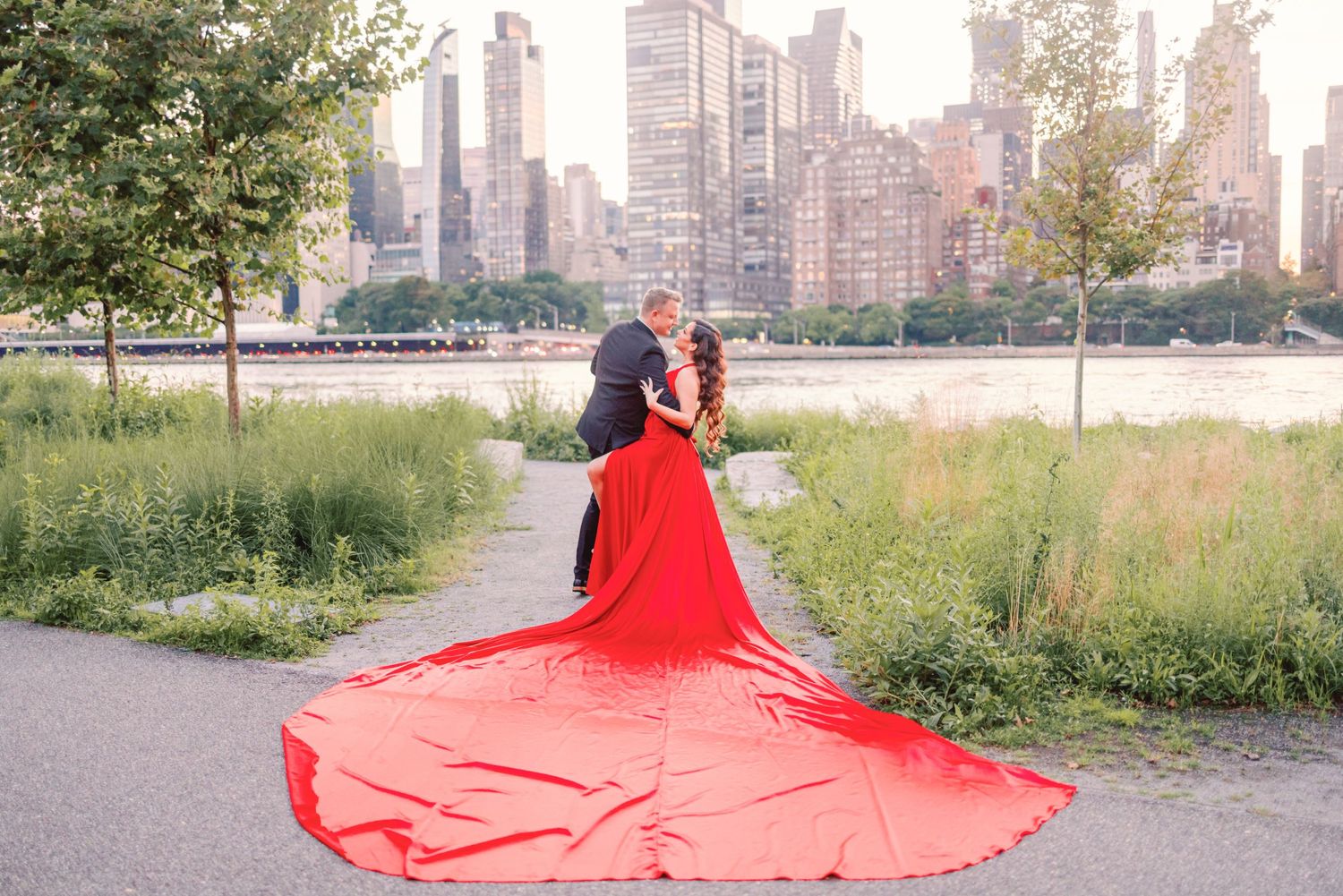 Couple sharing a romantic kiss in a stunning red wedding gown by the NYC skyline waterfront.