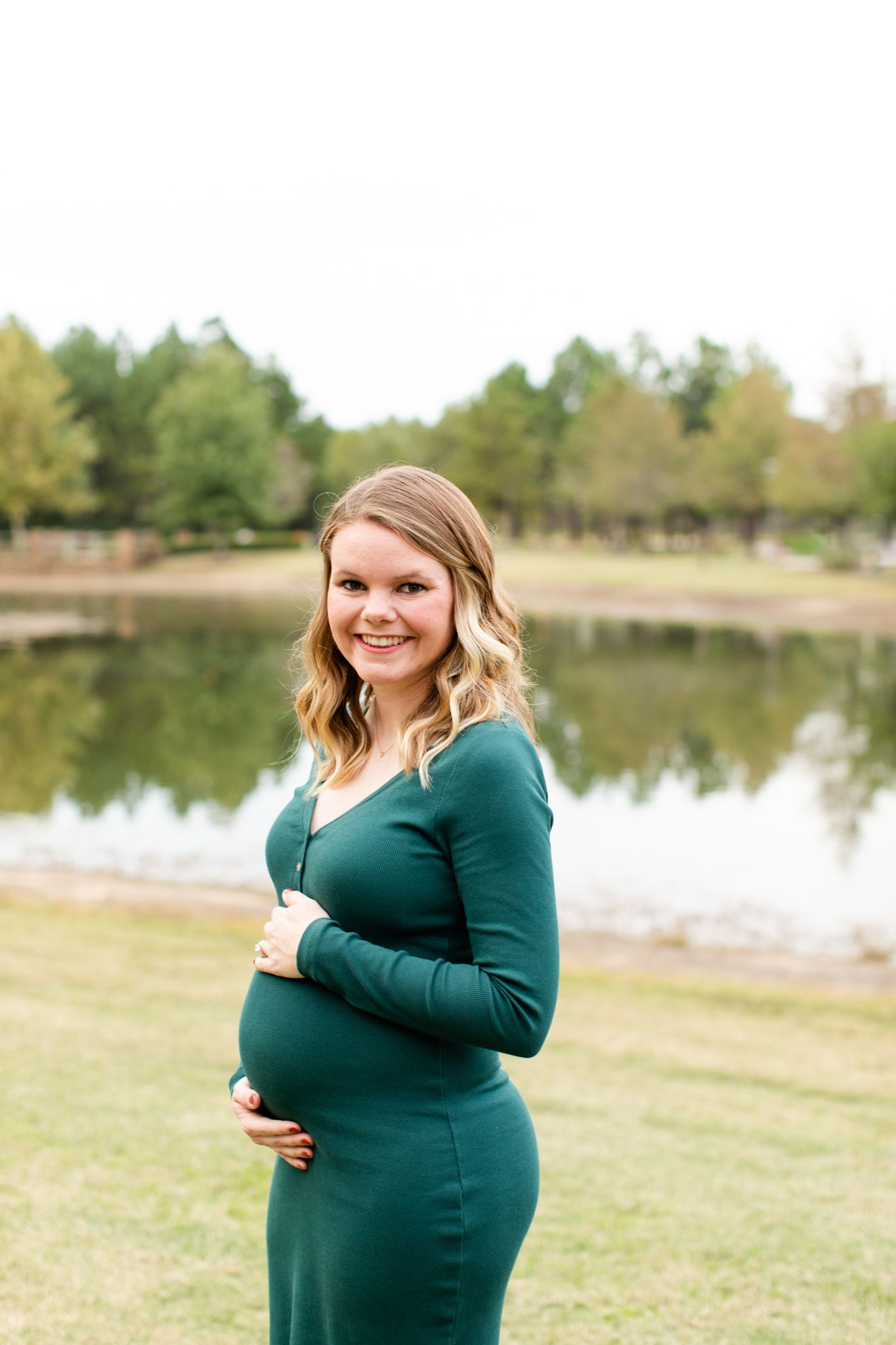 Expectant mother in green dress poses by lake during maternity photoshoot.