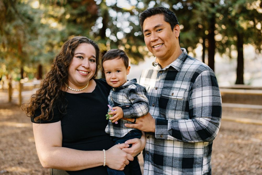 Family of three posing outdoors in casual attire against a natural forest backdrop.