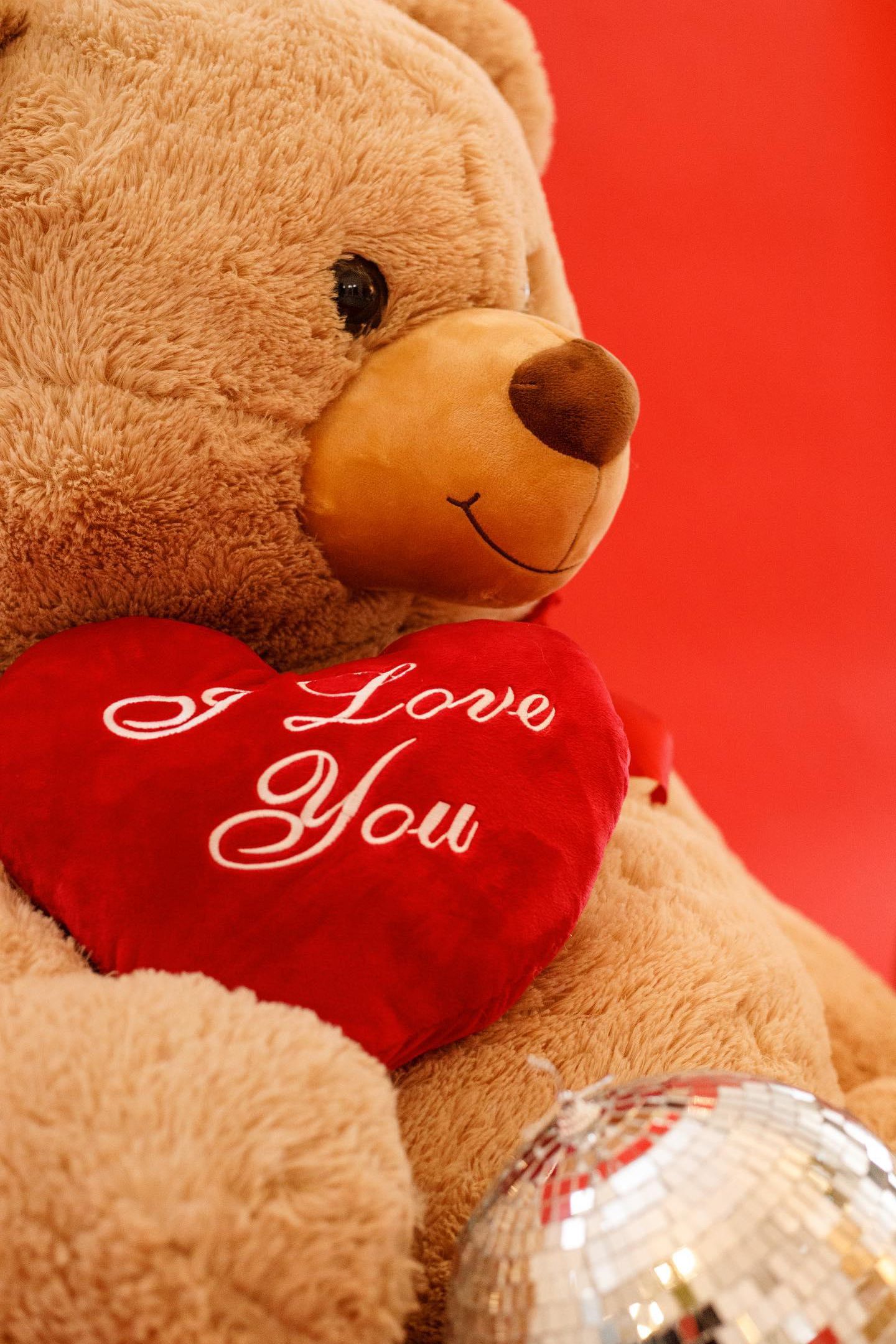Close-up of teddy bear holding red 'I Love You' heart pillow against red background.