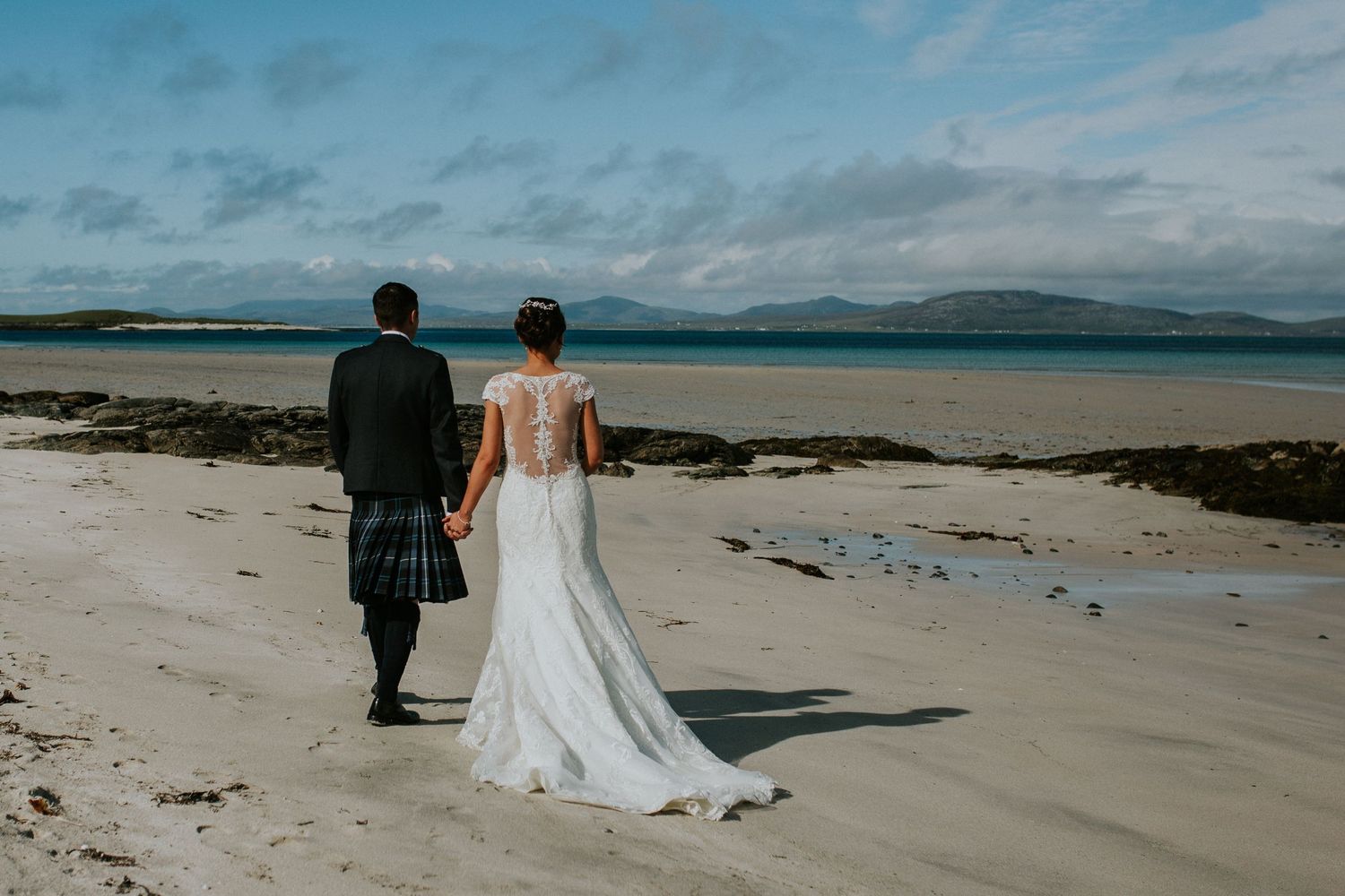 Wedding couple walking on beach with Scottish kilt and lace wedding dress.