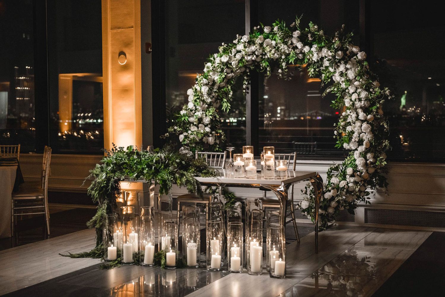 Romantic wedding reception setup with floral arch and glowing candles at night.