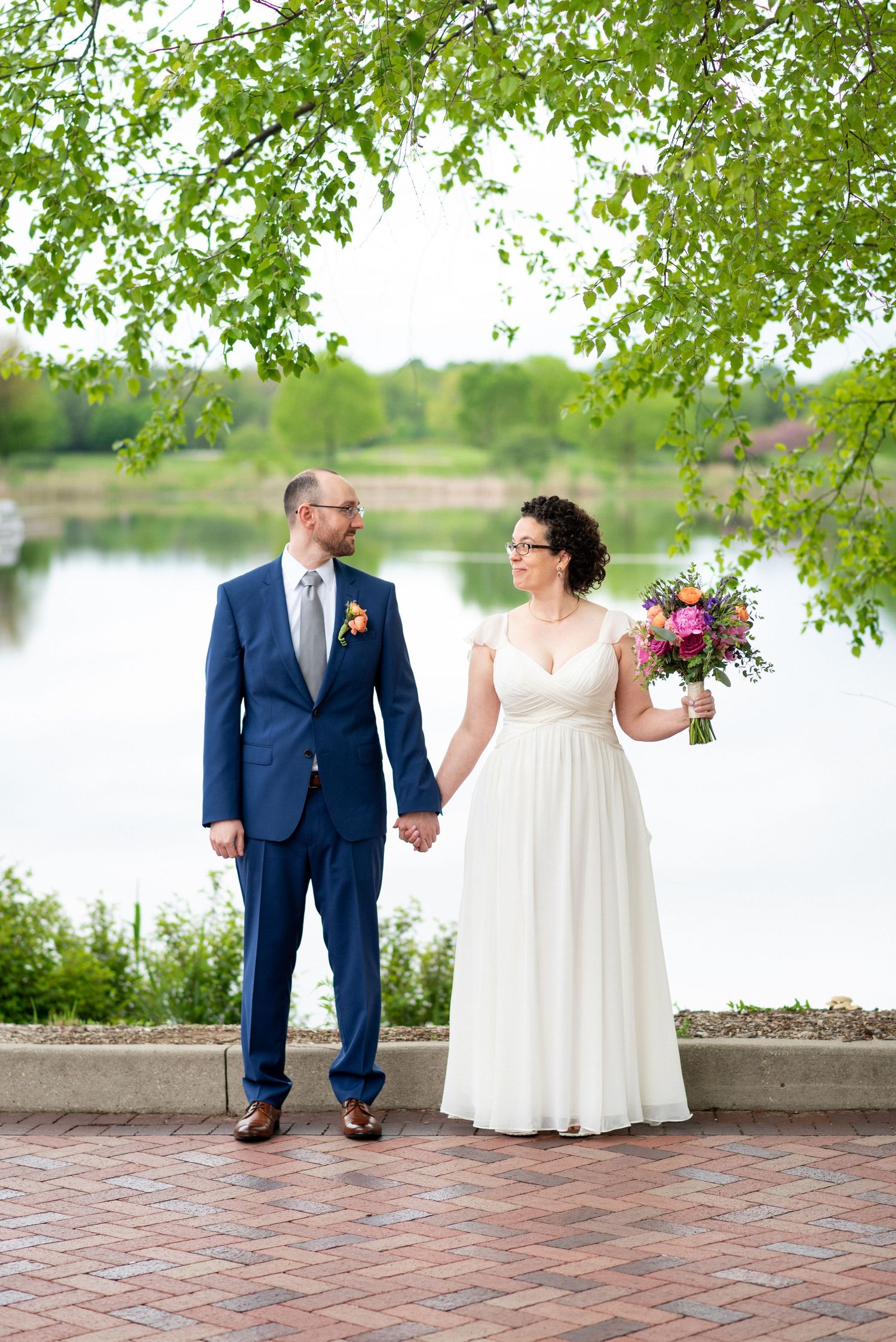 Bride and groom holding hands looking at eachother standing infront of a pond with green trees above and in backgroud.