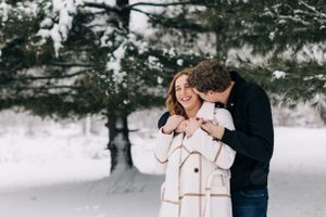 Couple embracing and laughing in a snowy winter forest setting with evergreen trees.