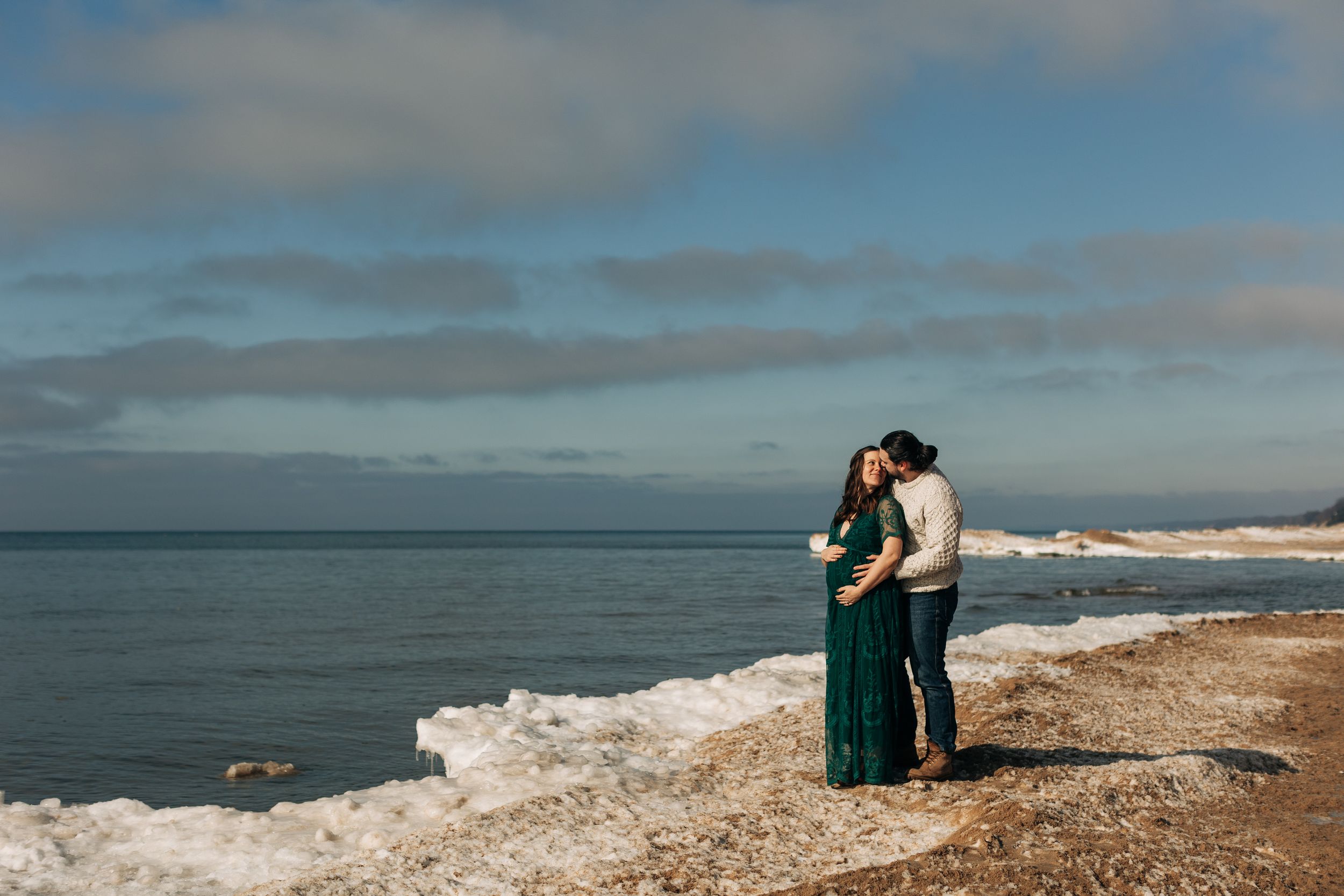 Couple embracing on rocky coastline with ocean waves and blue sky backdrop.