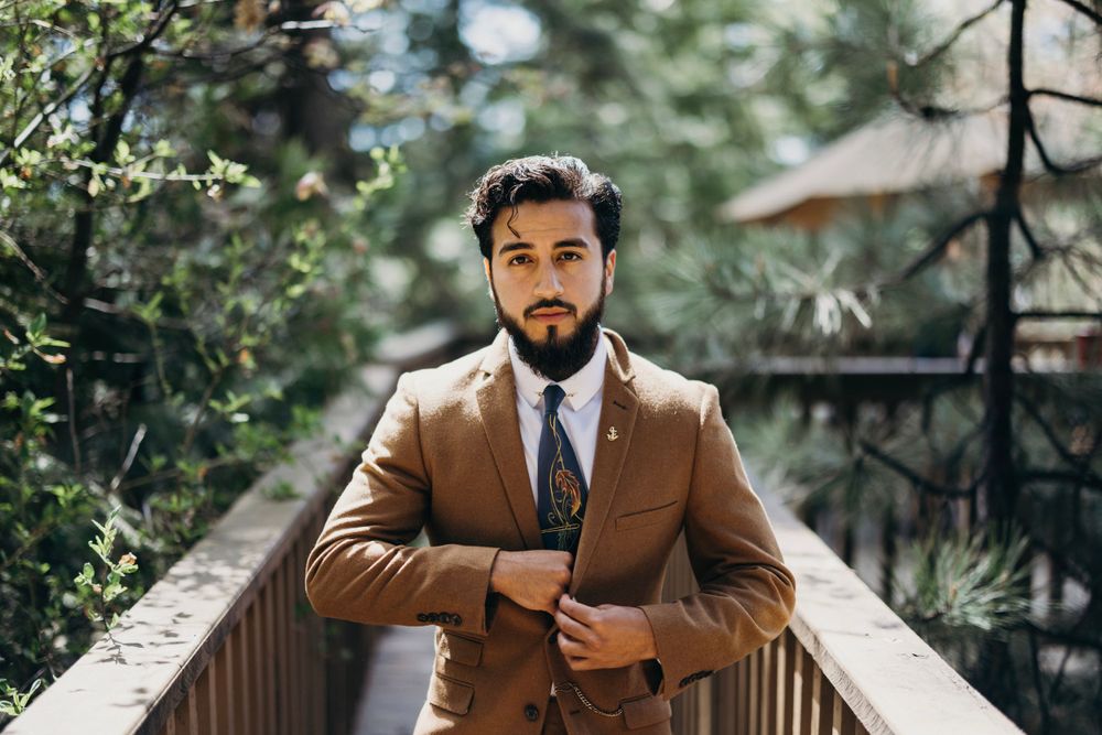 Stylish groom in brown suit poses outdoors against lush green foliage.