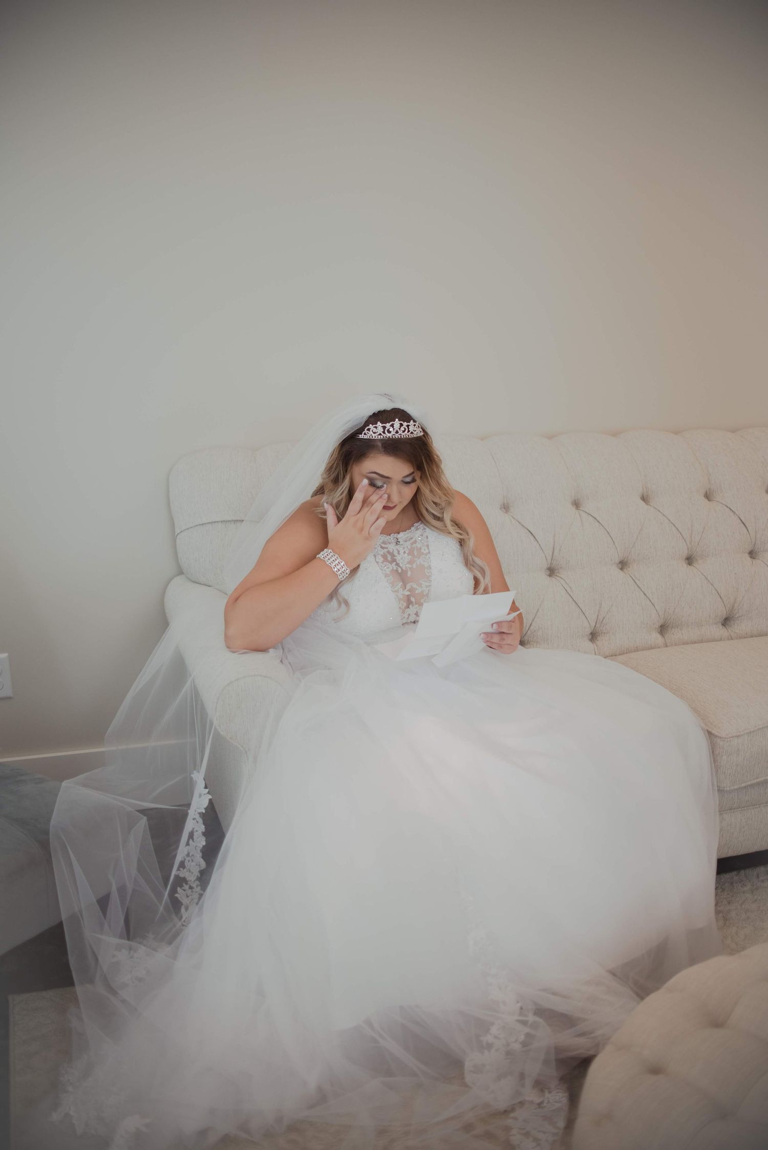 Bride in flowing white gown reading emotional letter while seated on tufted sofa.