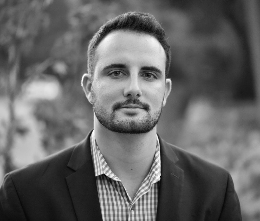 Professional headshot of man wearing blazer and striped shirt against blurred background.