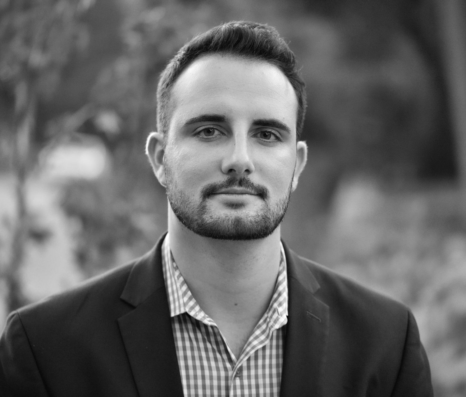 Professional headshot of man wearing blazer and striped shirt against blurred background.