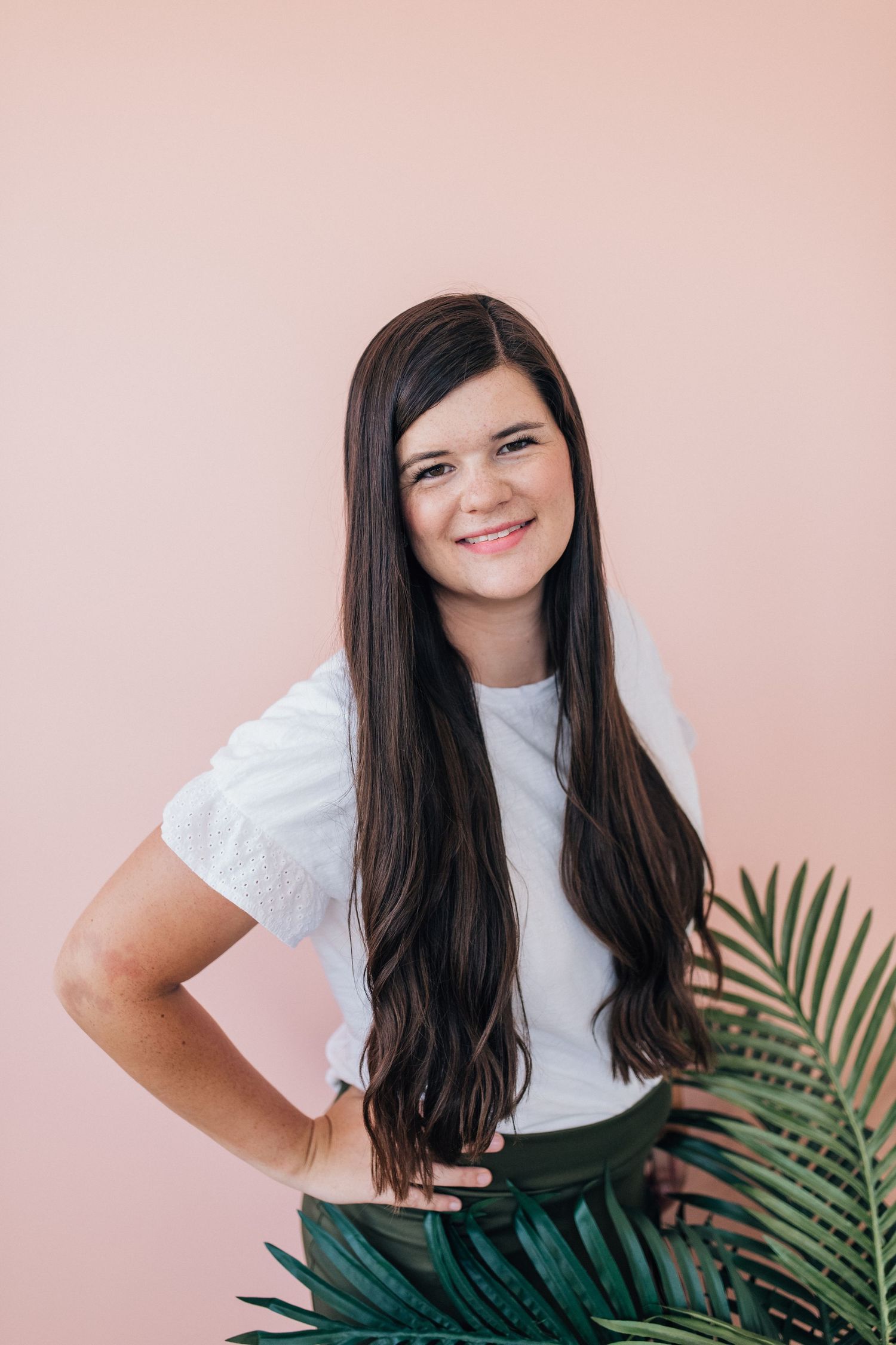 A person in a white shirt stands against a pink wall with tropical plants.