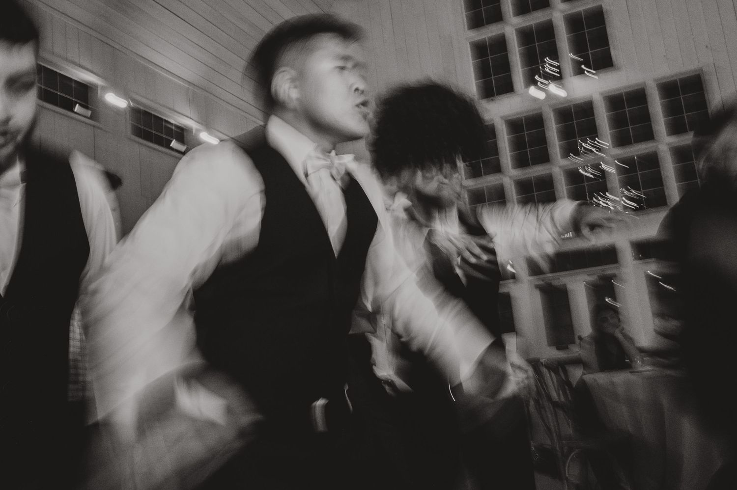 Motion-blurred black and white photo of wedding guests dancing at a lively reception.