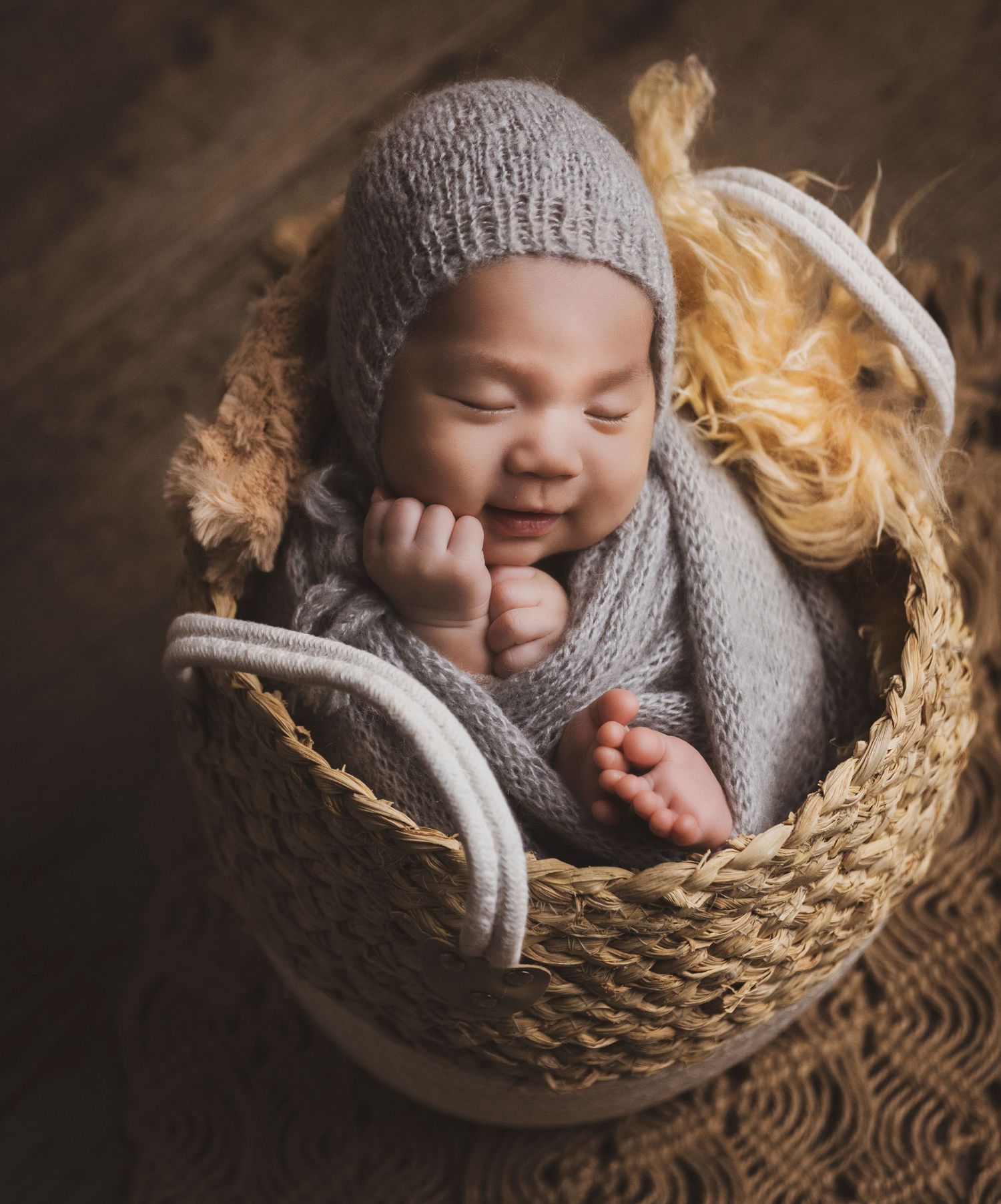 Sleeping newborn in gray knit wrap and bonnet hat posed in a wicker basket with fluffy accents.