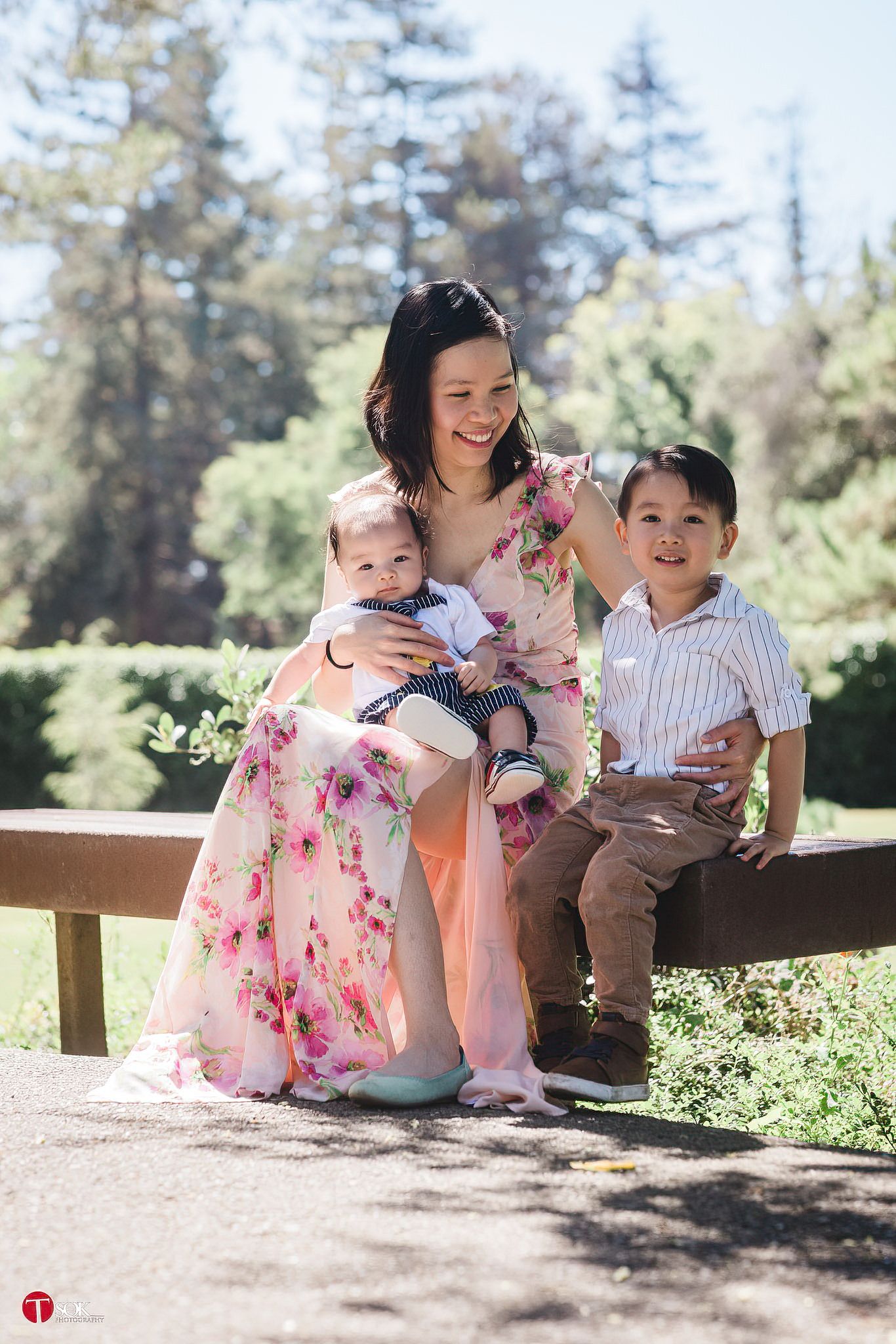 A woman in a floral dress sits with children on a wooden bench in a park.