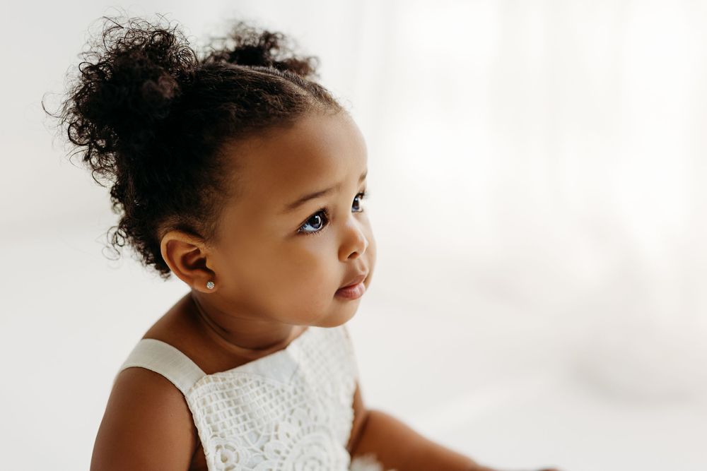 A young child wearing a white eyelet dress gazes thoughtfully to the side against a light background. during a session in Fairfax, VA.