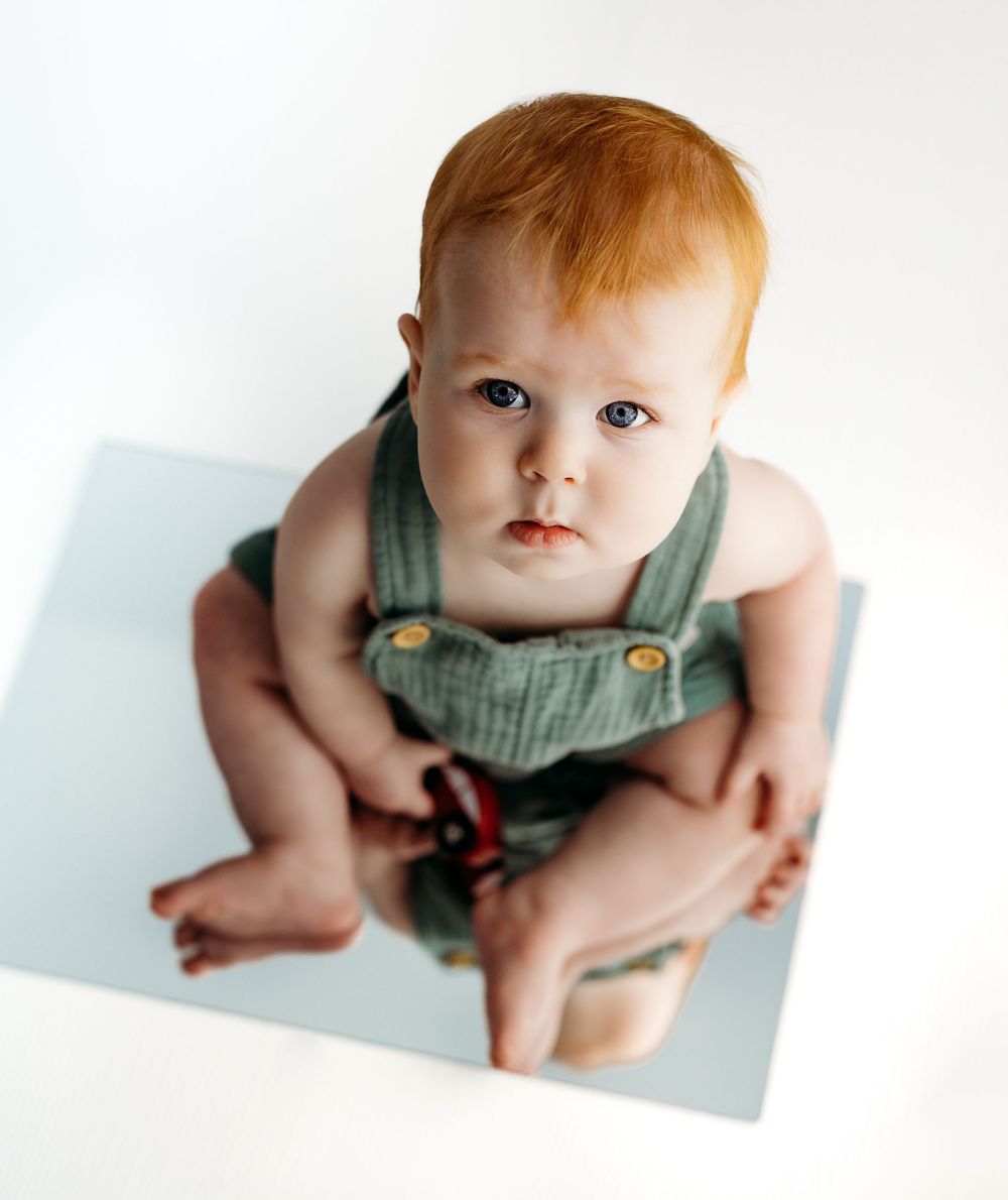 Toddler in green overalls sits looking at camera against white background. during a session in Fairfax, VA.